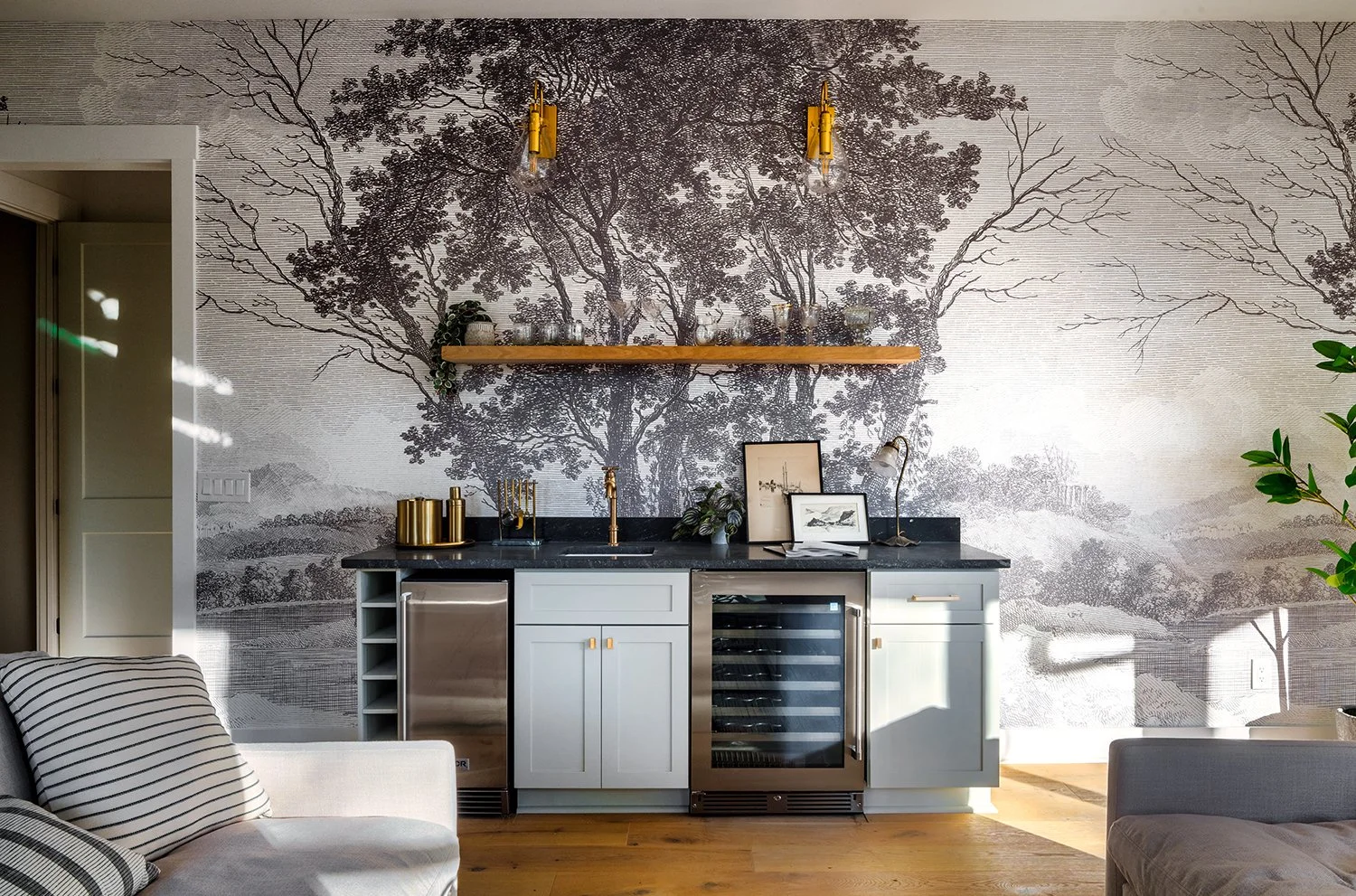 Interior home bar area with a black countertop, light blue cabinets, and a wine cooler. A wooden shelf above holds decorative items and glassware. The wall features a large black-and-white mural of trees and landscape, with shelves and framed art leaning against the wall. Light-colored armchairs with striped pillows are partially visible in the foreground.