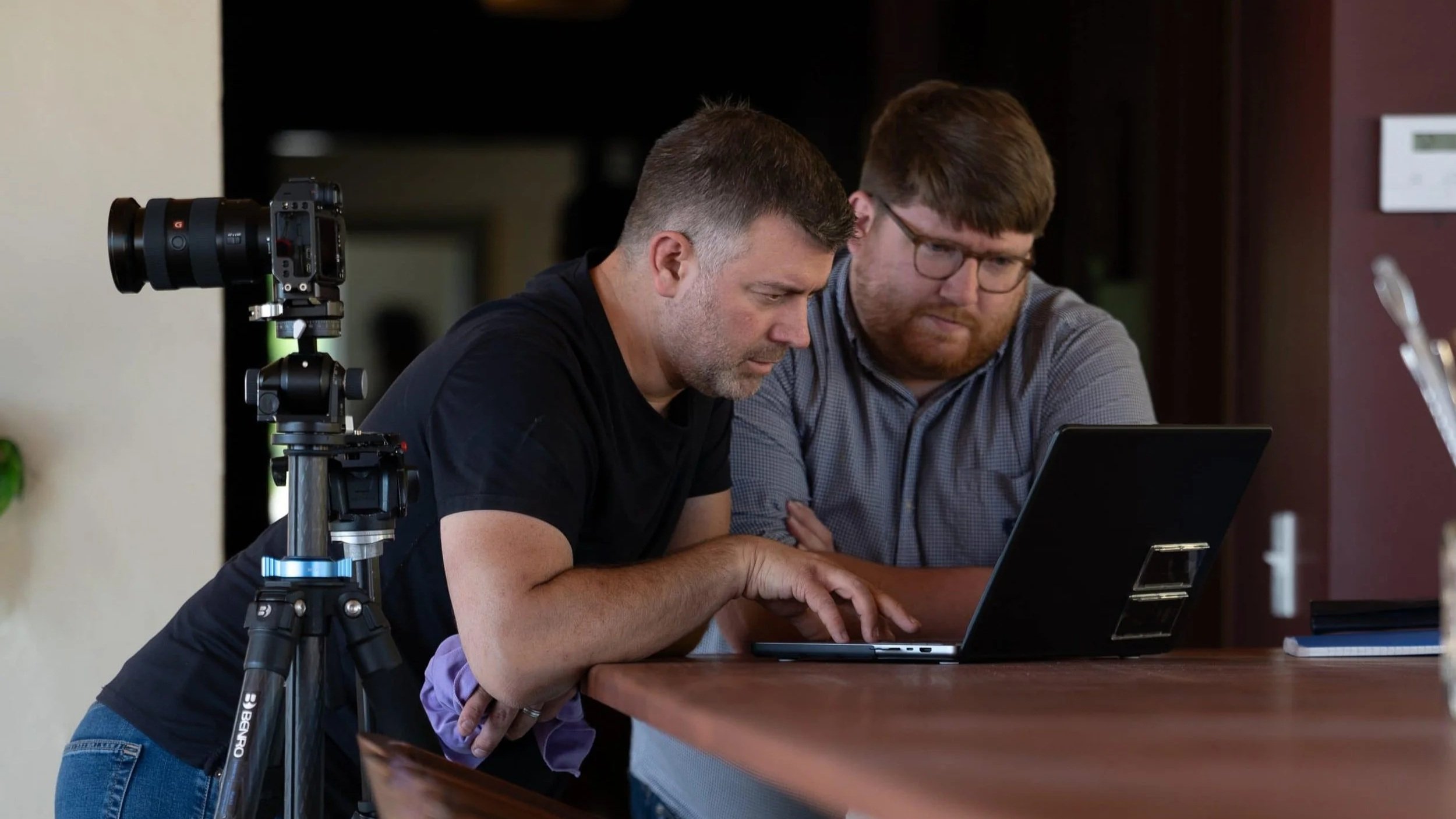 Two men look at a laptop screen together at a wooden table. One wears a black shirt, the other glasses and a checkered shirt. A camera on a tripod is positioned to the side.