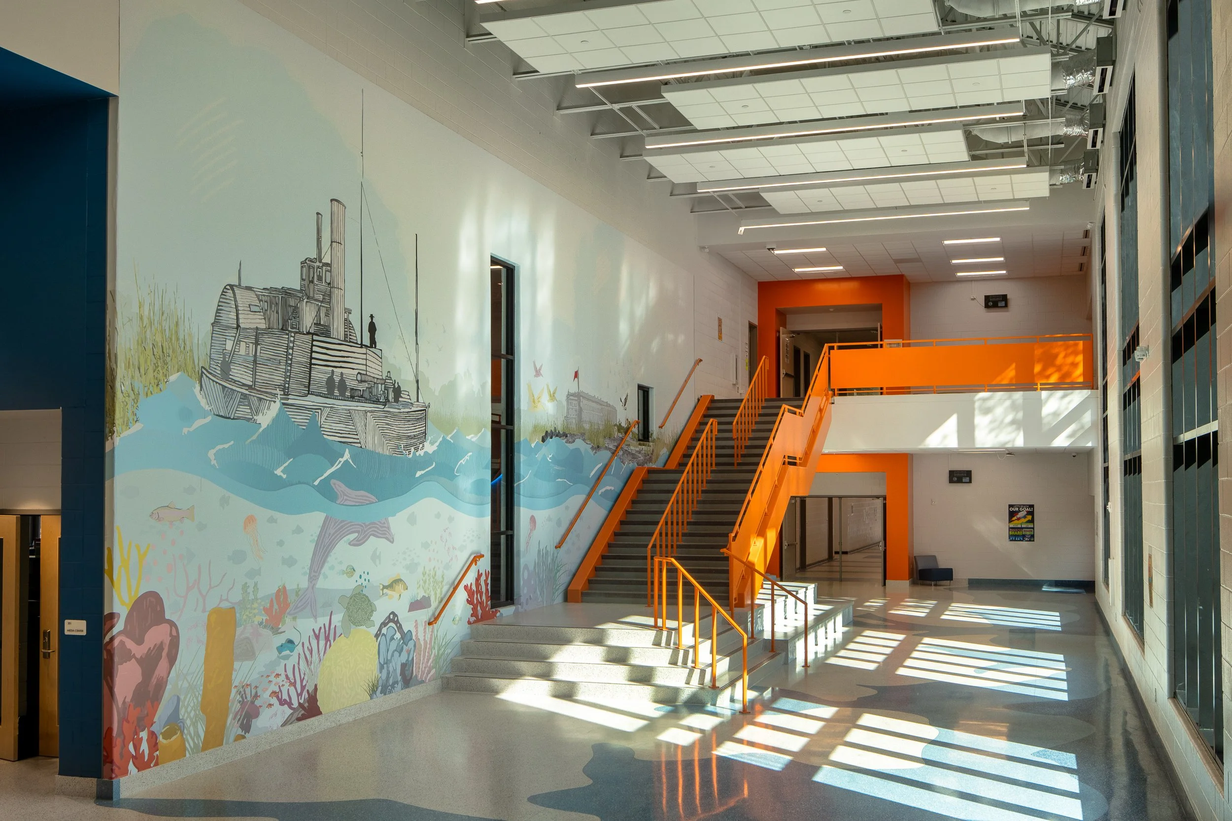 Tall school stair hall with orange railings, colorful coastal wall graphics, overhead acoustic panels, and sunlight across the floor.