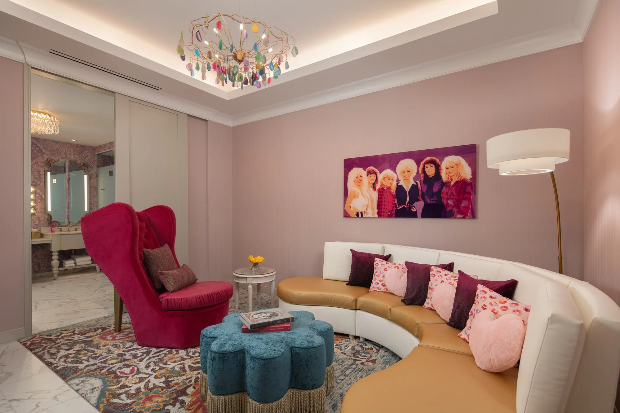 Colorful hotel sitting room in the Six Sisters Suite with a curved white and gold sofa, sculptural pink chair, blue ottoman, artwork, and chandelier.