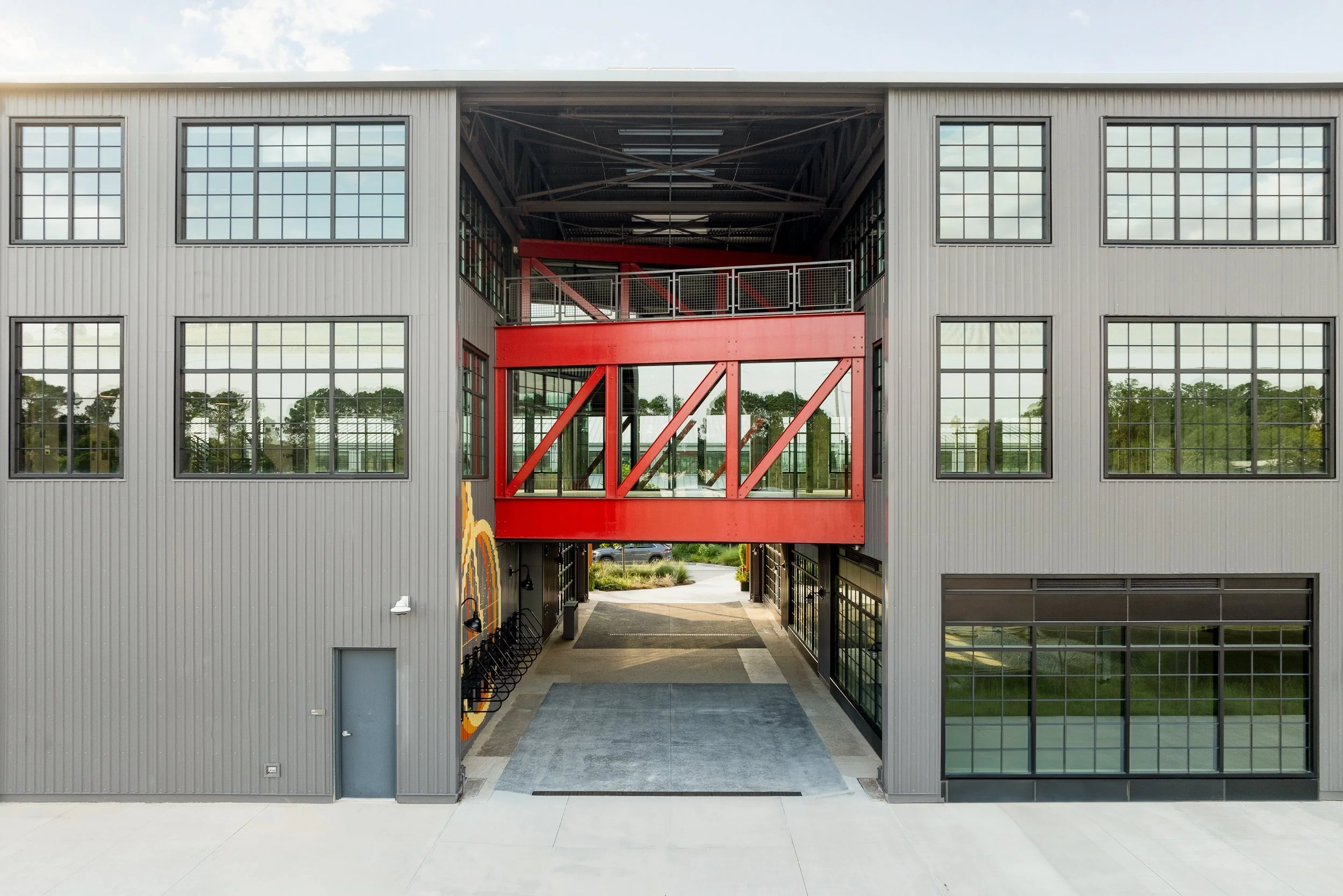 Symmetrical view of the central breezeway with a vibrant red diagonal-braced steel bridge spanning the second level, industrial-style windows on either side, and a concrete drive-through below.