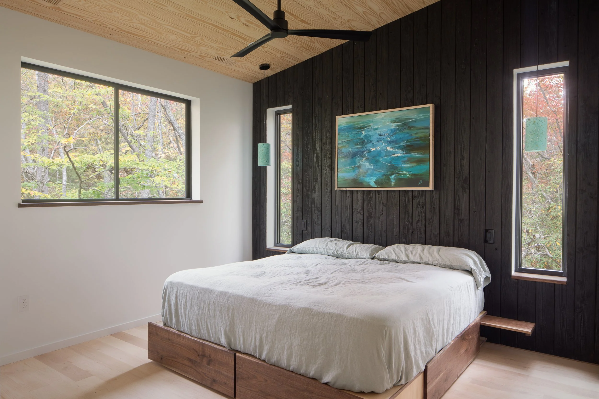 Bedroom with a burnt cedar accent wall, low platform bed, wood ceiling, and tall windows framing the trees.