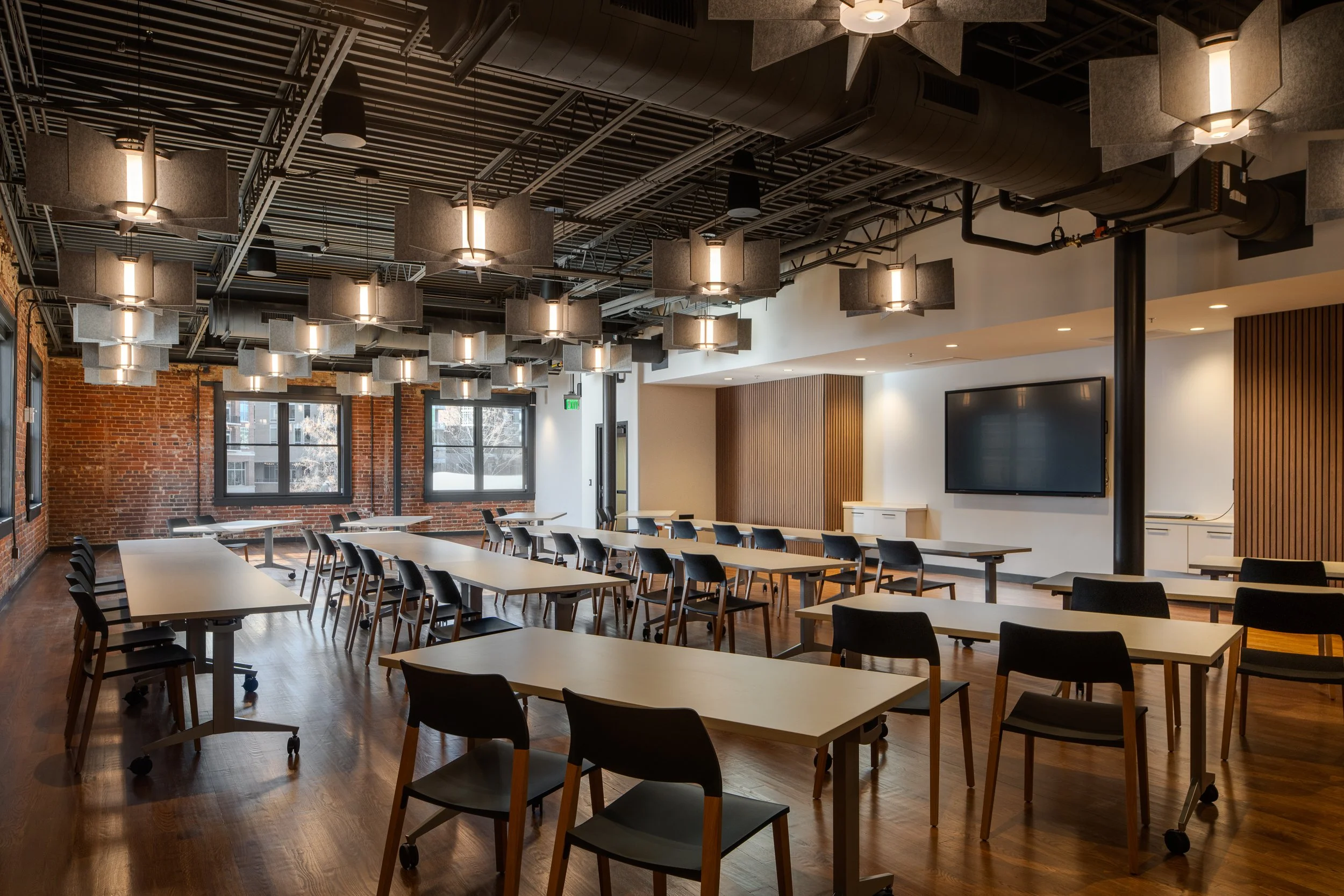Classroom-style meeting room with rows of light wood tables and black chairs, exposed brick walls, and hexagonal acoustic baffles suspended from the open ceiling.