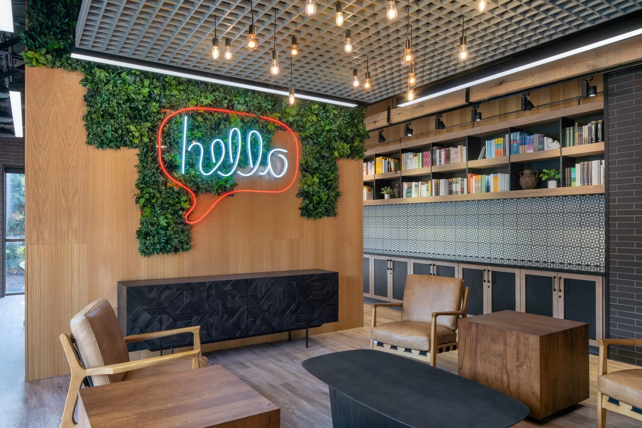Office lounge with leather chairs, wood millwork, a green plant wall, bookshelves, patterned tile, and a neon hello sign.