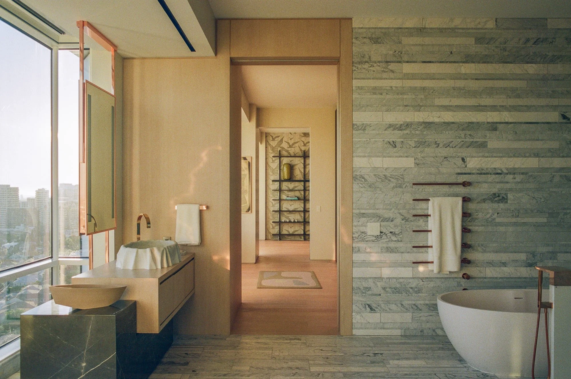 View through a primary bath with stone tile, freestanding tub, floating vanity, and warm afternoon light.