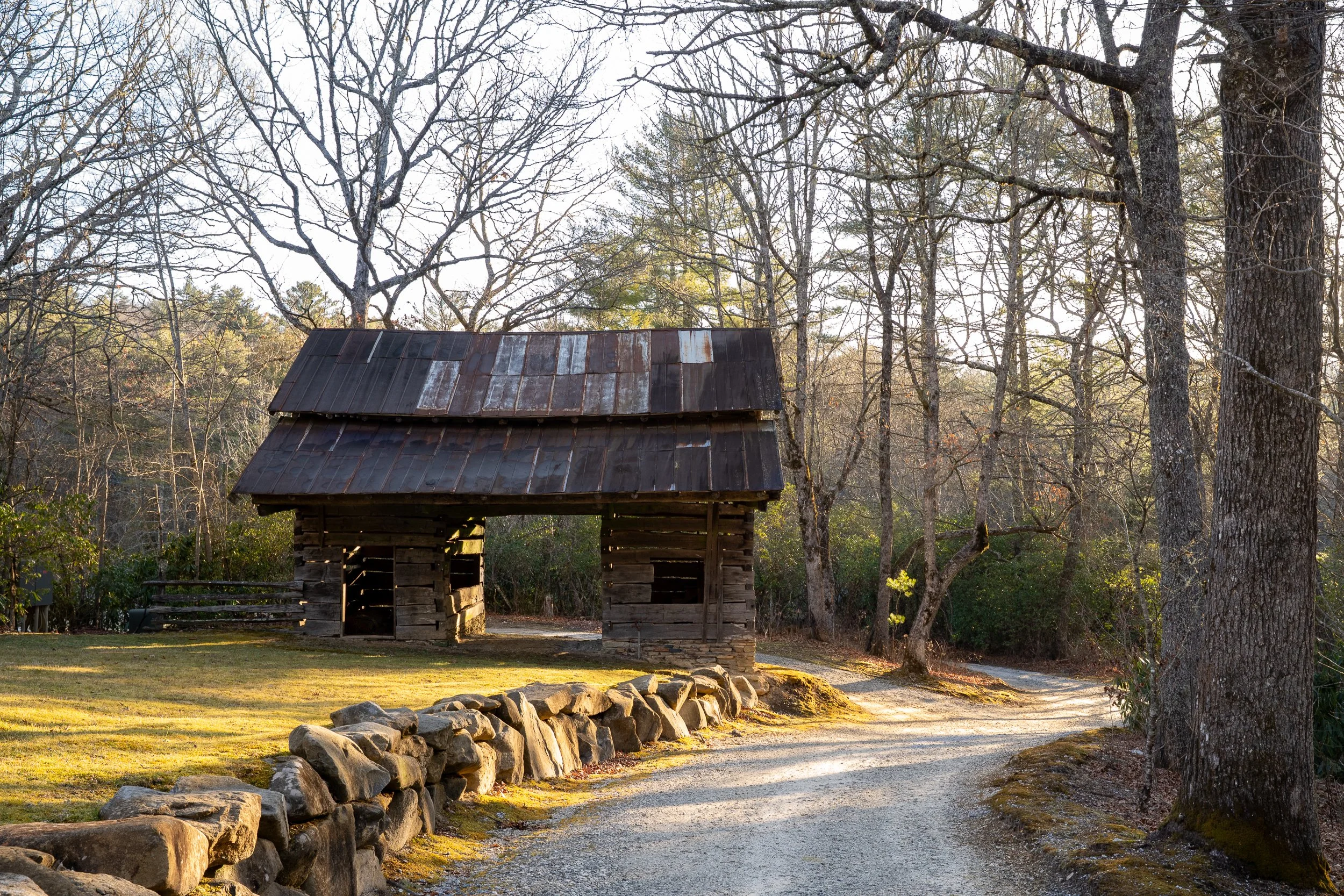 Weathered barn structure beside a gravel road, stone wall, and bare winter trees.