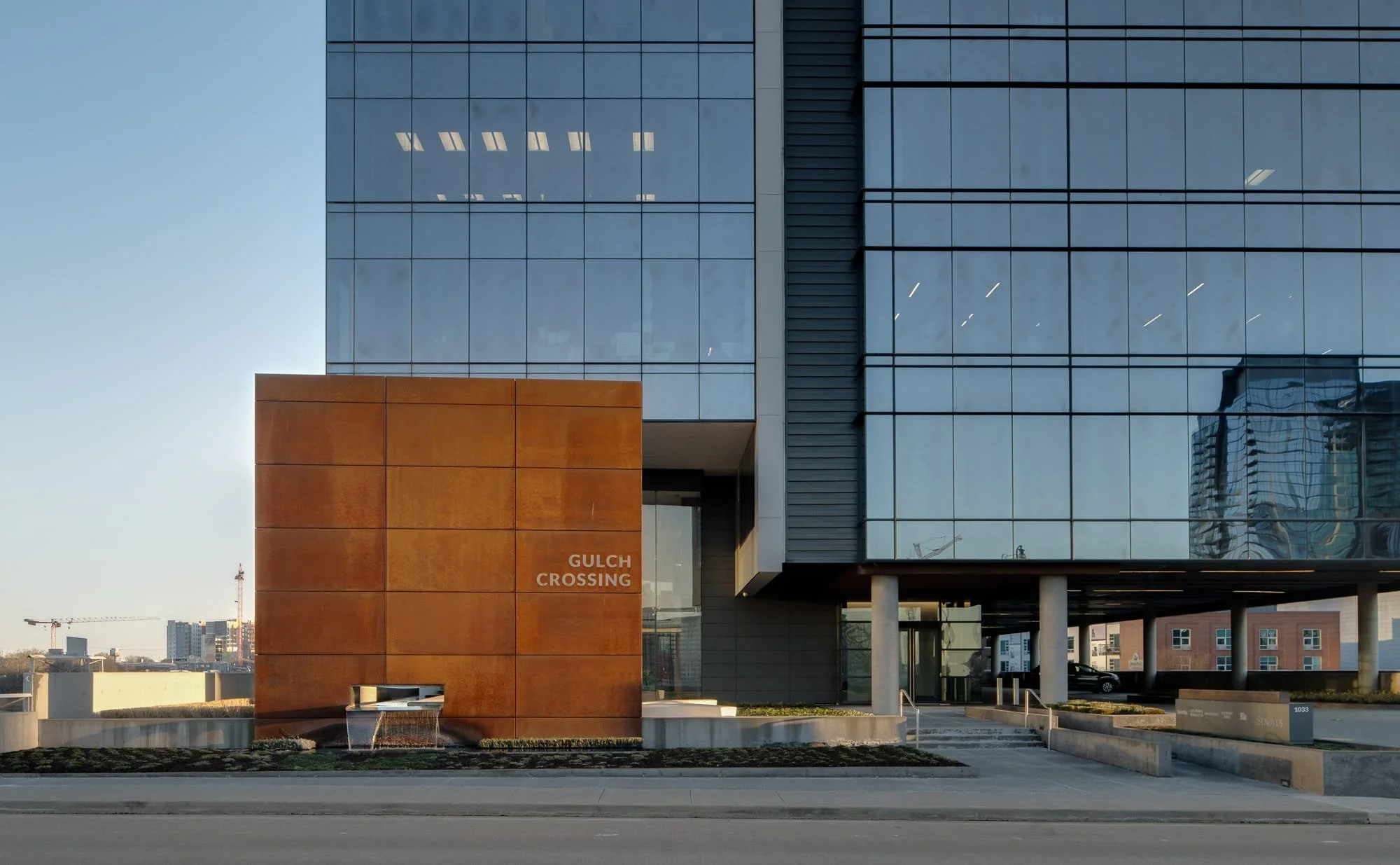 Street level view of Gulch Crossing in Nashville showing the corten steel entry cube with building signage, curtain wall glazing, and a construction crane visible in the distance