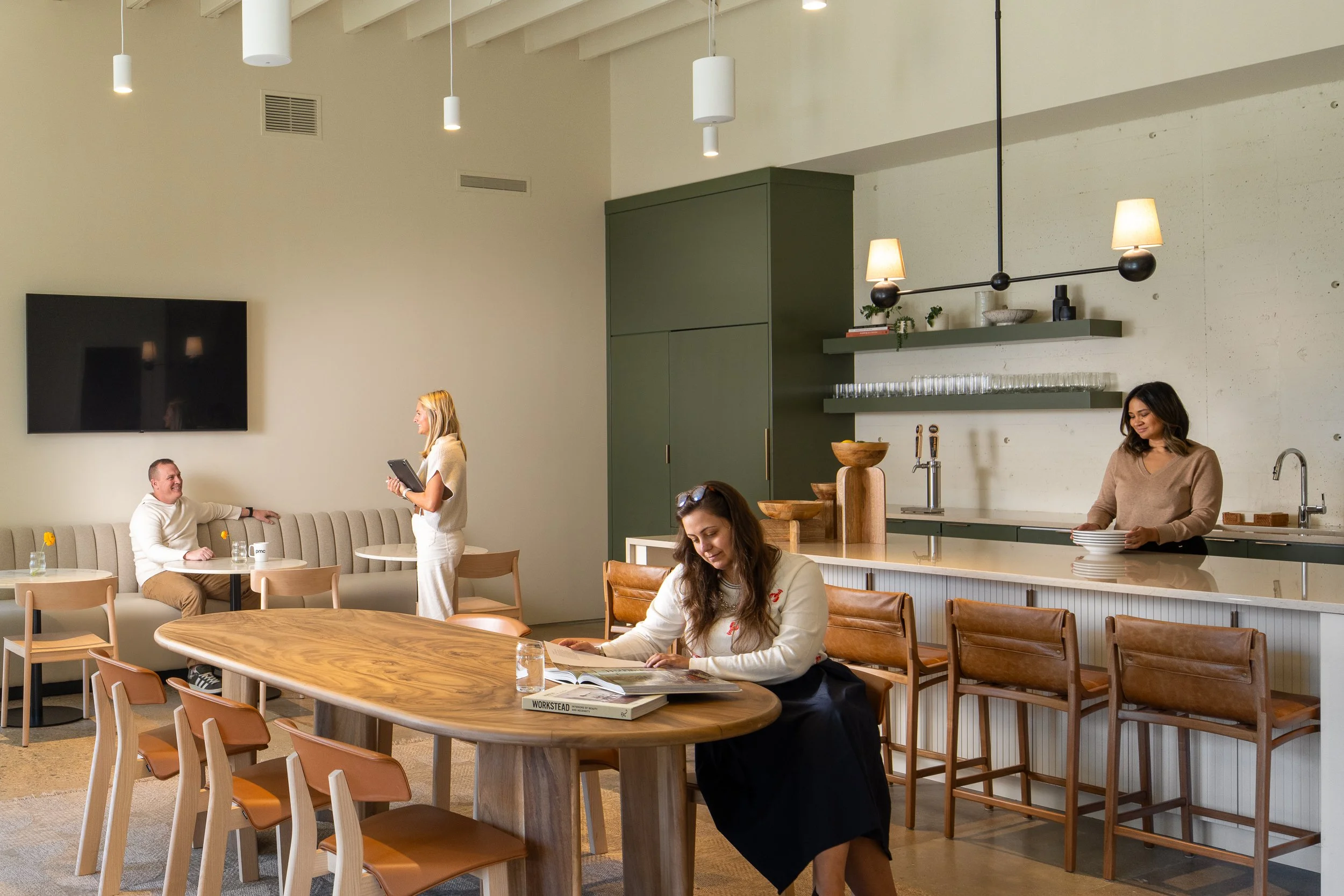 Open cafe area with a green millwork tap counter, white quartz island, oval wood dining table, leather counter stools, and a tan banquette where staff gather for coffee and conversation.