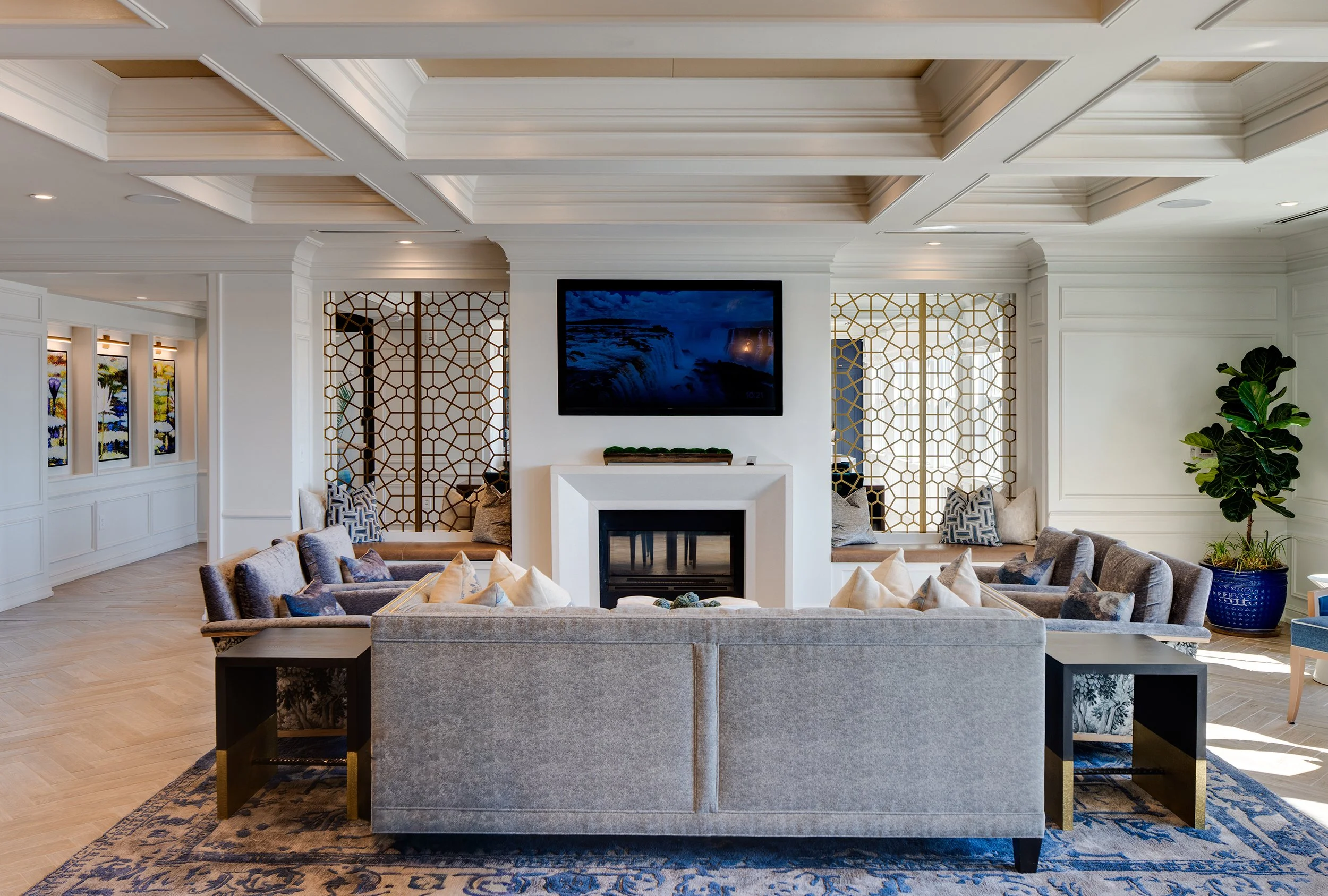 Resident lounge with a deep coffered ceiling, central fireplace beneath a mounted screen, paired window seats flanked by gold geometric screens, gray velvet sofas, and a blue Persian-style rug.