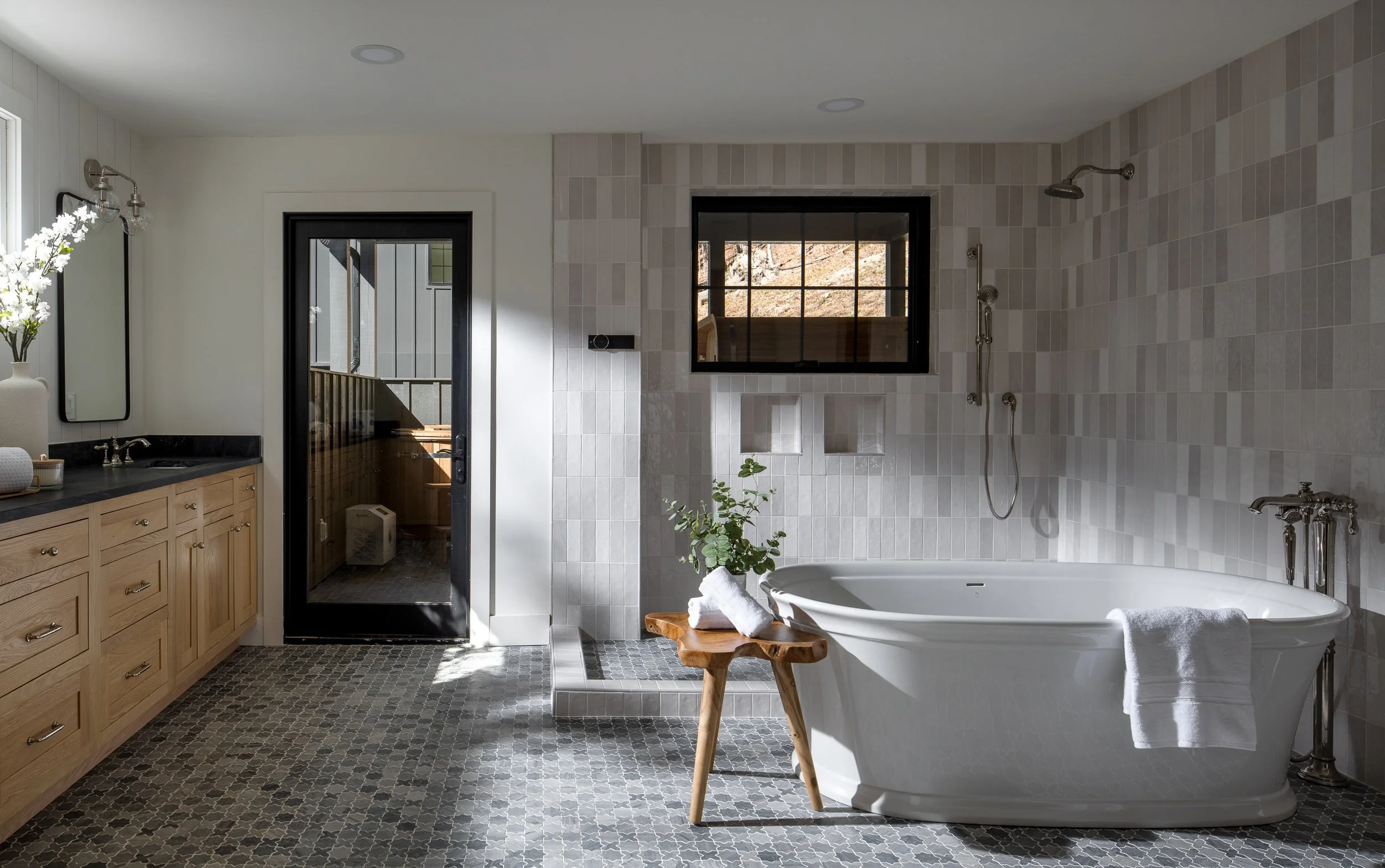 Primary bathroom with freestanding tub, tiled shower wall, wood vanity, black-framed door, and patterned floor tile.