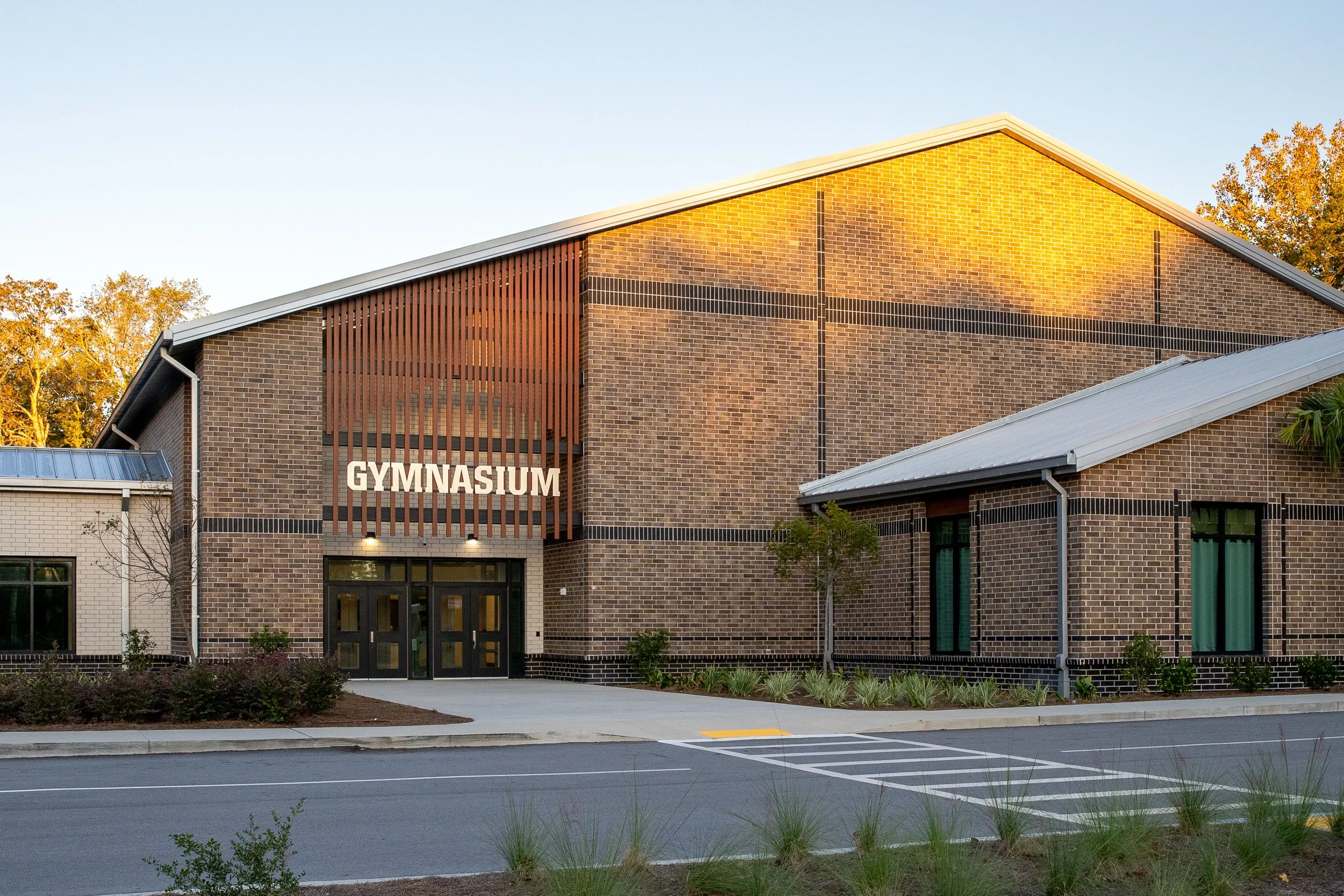 Gymnasium entrance with brick facade, metal roof, wood screen detail, and warm golden-hour light across the exterior wall.