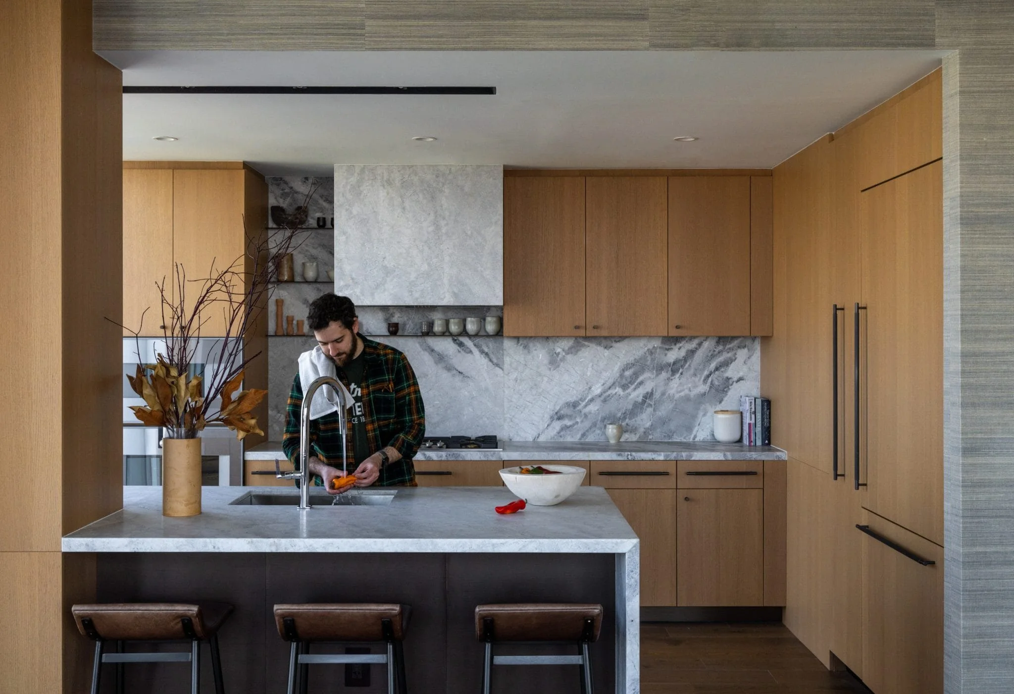 Modern residential kitchen with light wood cabinetry, marble backsplash, island seating, and a person preparing food at the sink.