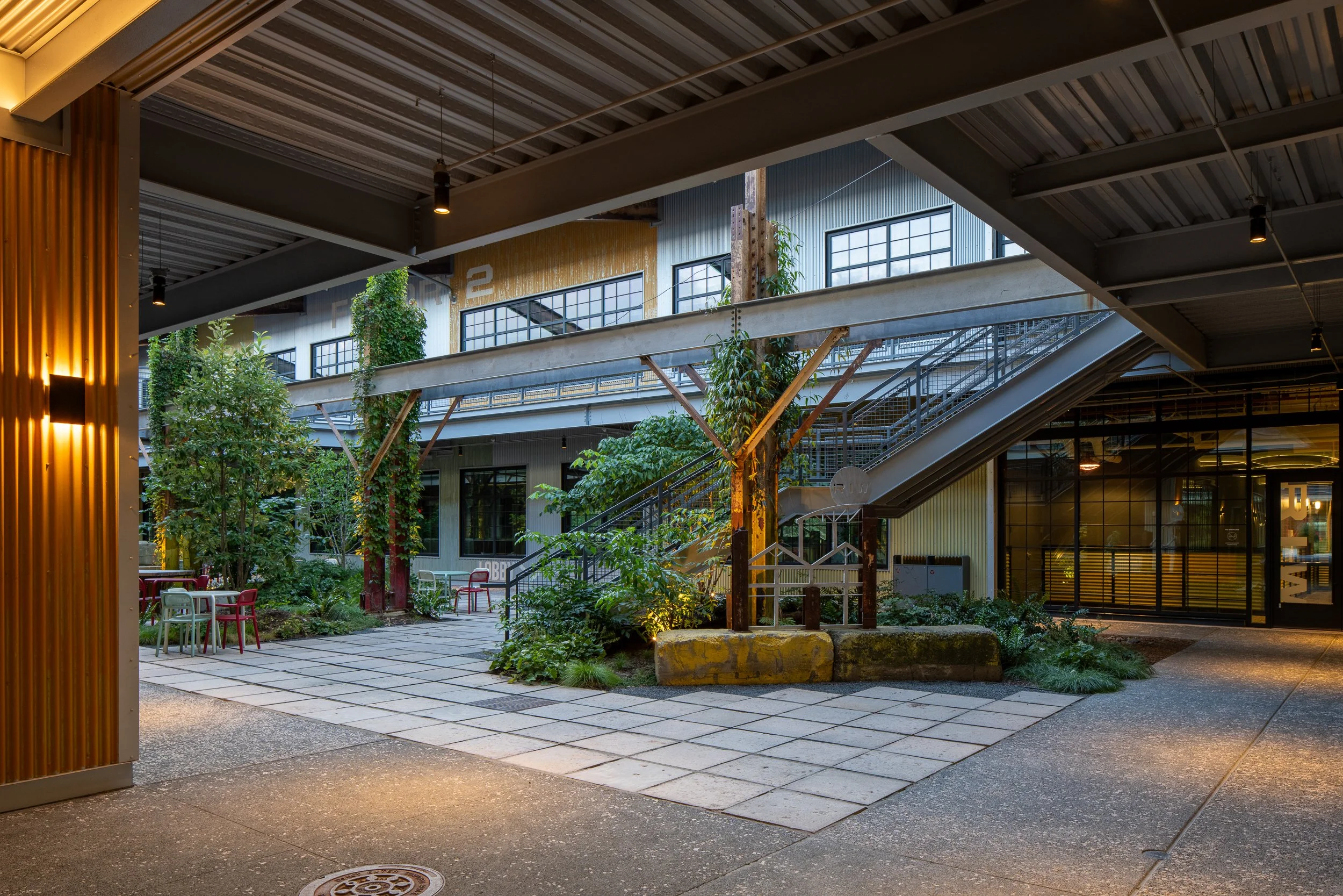 Interior pedestrian plaza beneath a metal canopy with a rusted steel column wrapped in climbing vines, mint and red bistro chairs, and the lit RIW gateway and elevated walkways beyond.