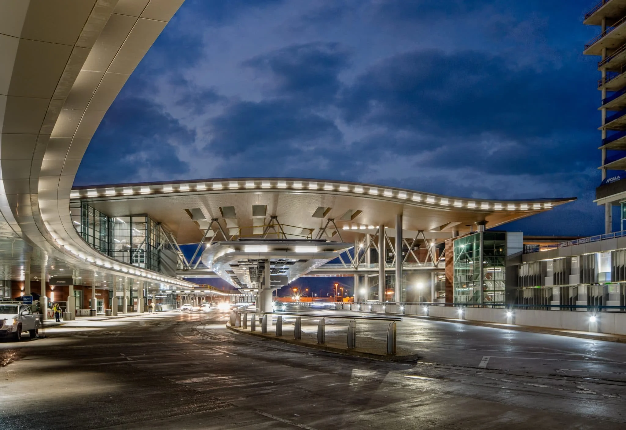 Exterior view of an airport drop-off area with wet pavement, dramatic evening clouds, terminal lighting, and a curved architectural canopy.