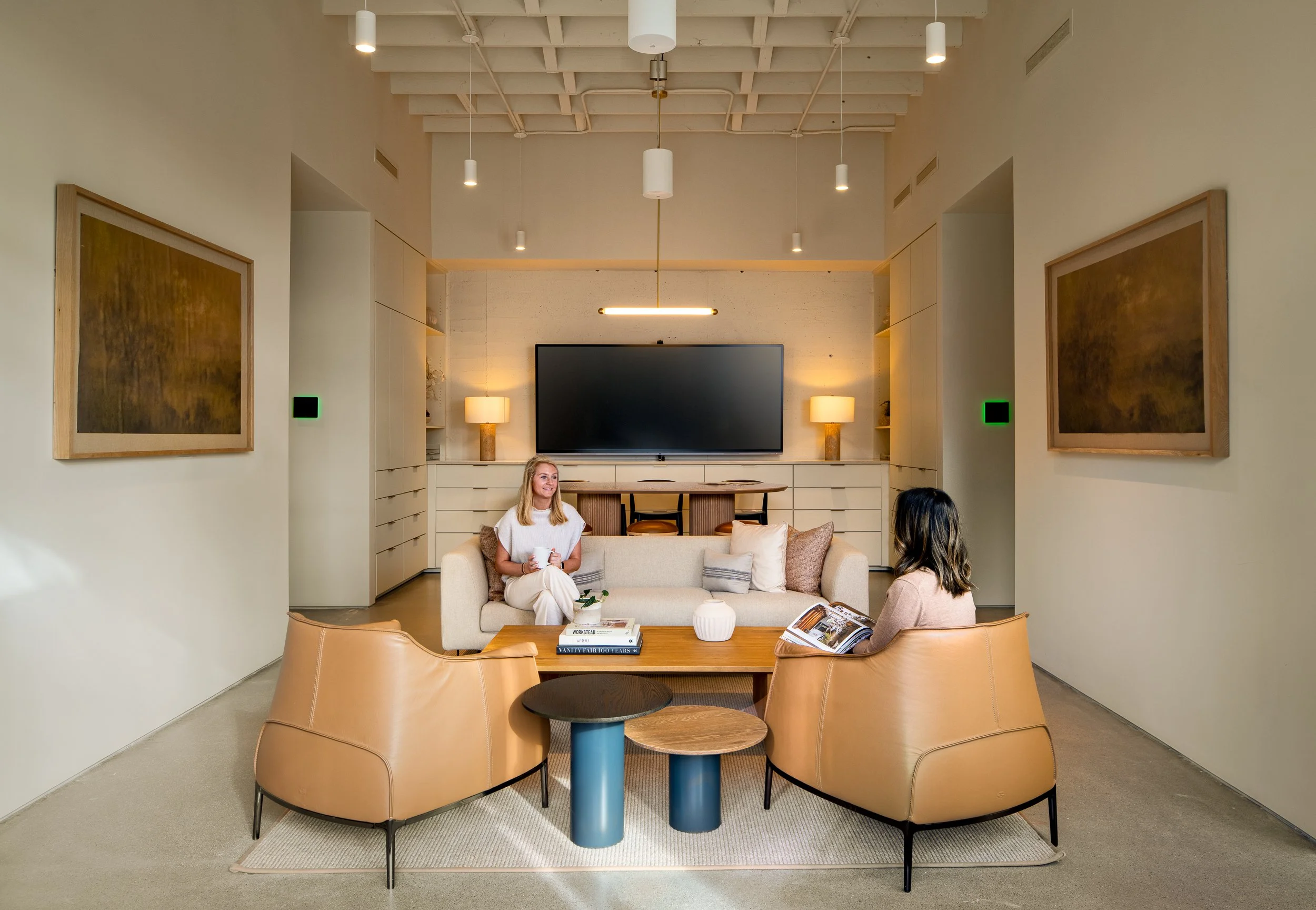 Interior conversation lounge with cognac leather chairs, a cream sofa, two abstract framed artworks, and a built-in display wall with a mounted screen flanked by lamp-lit shelving.