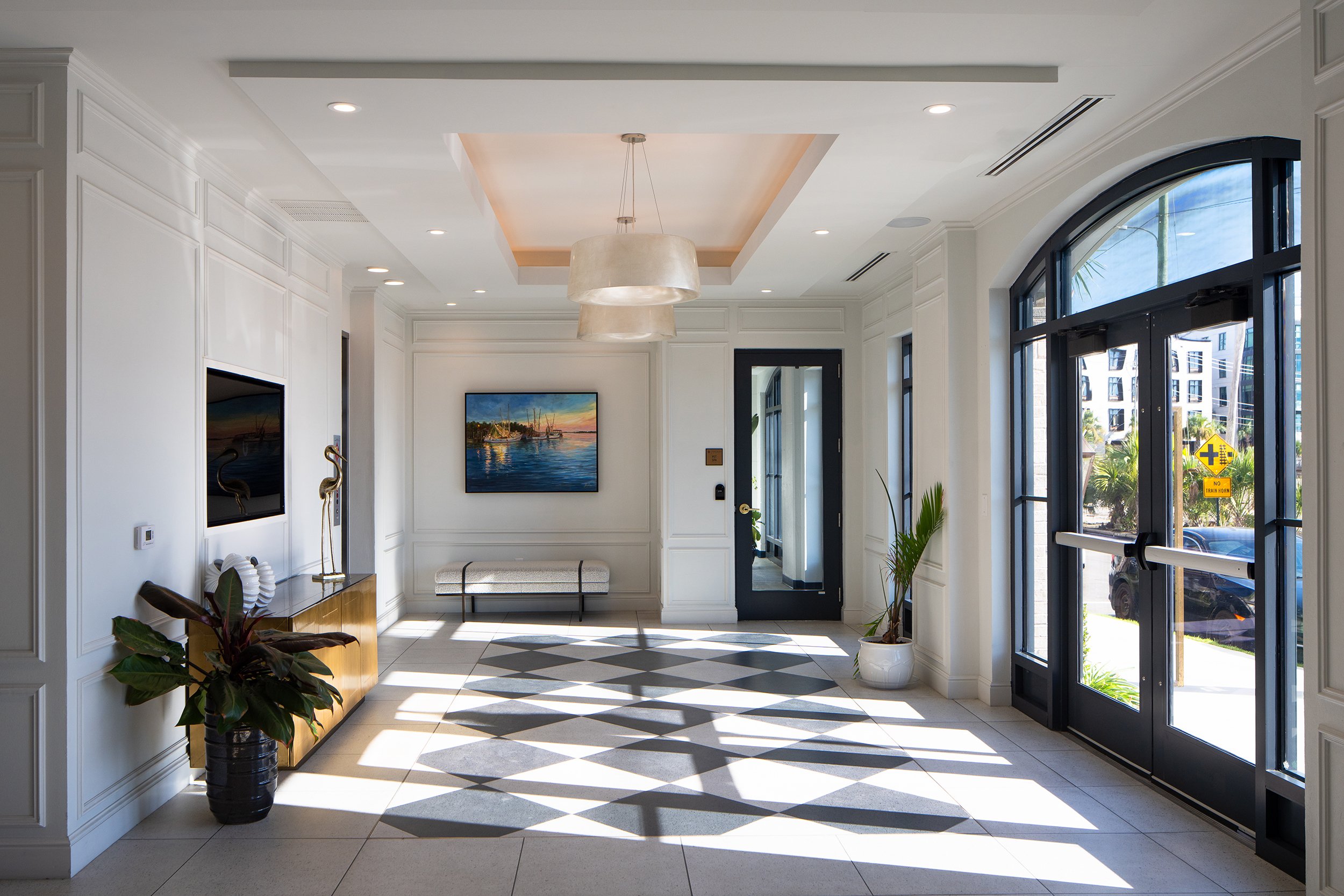 Side entry vestibule with a black and white harlequin diamond tile floor, paneled walls, a drum pendant, an upholstered bench, a framed Charleston harbor painting, and a paired arched glass door leading to the street.