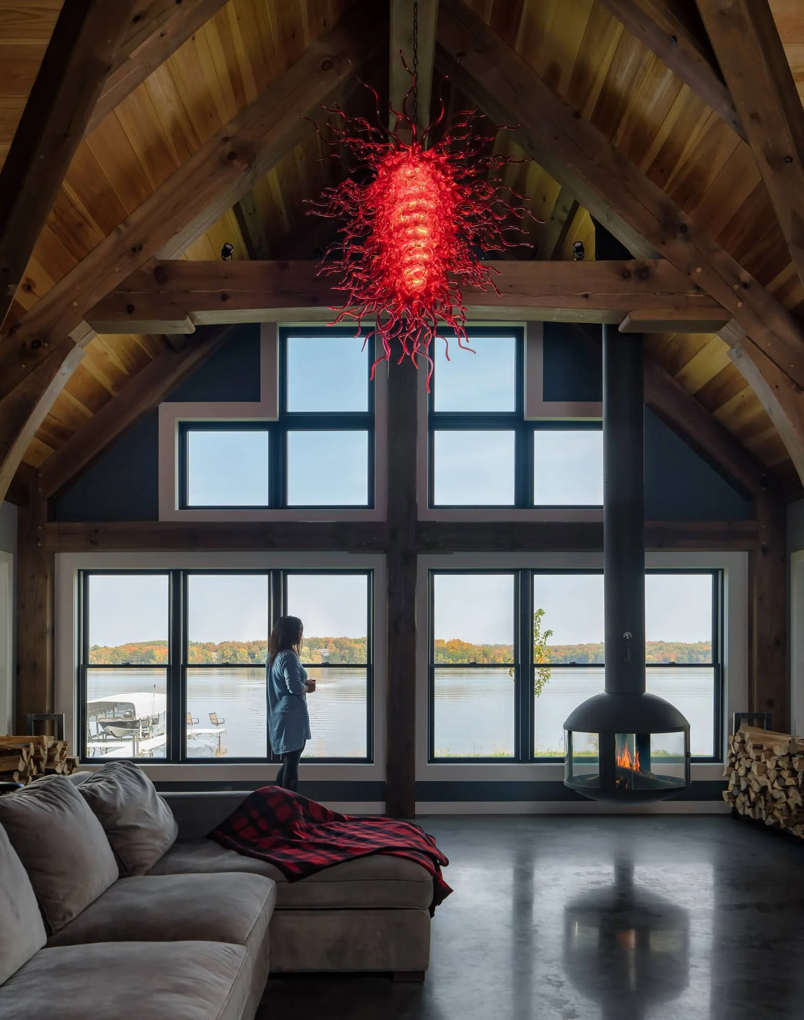 Vaulted living room with timber beams, lake-facing windows, red chandelier, and freestanding fireplace.