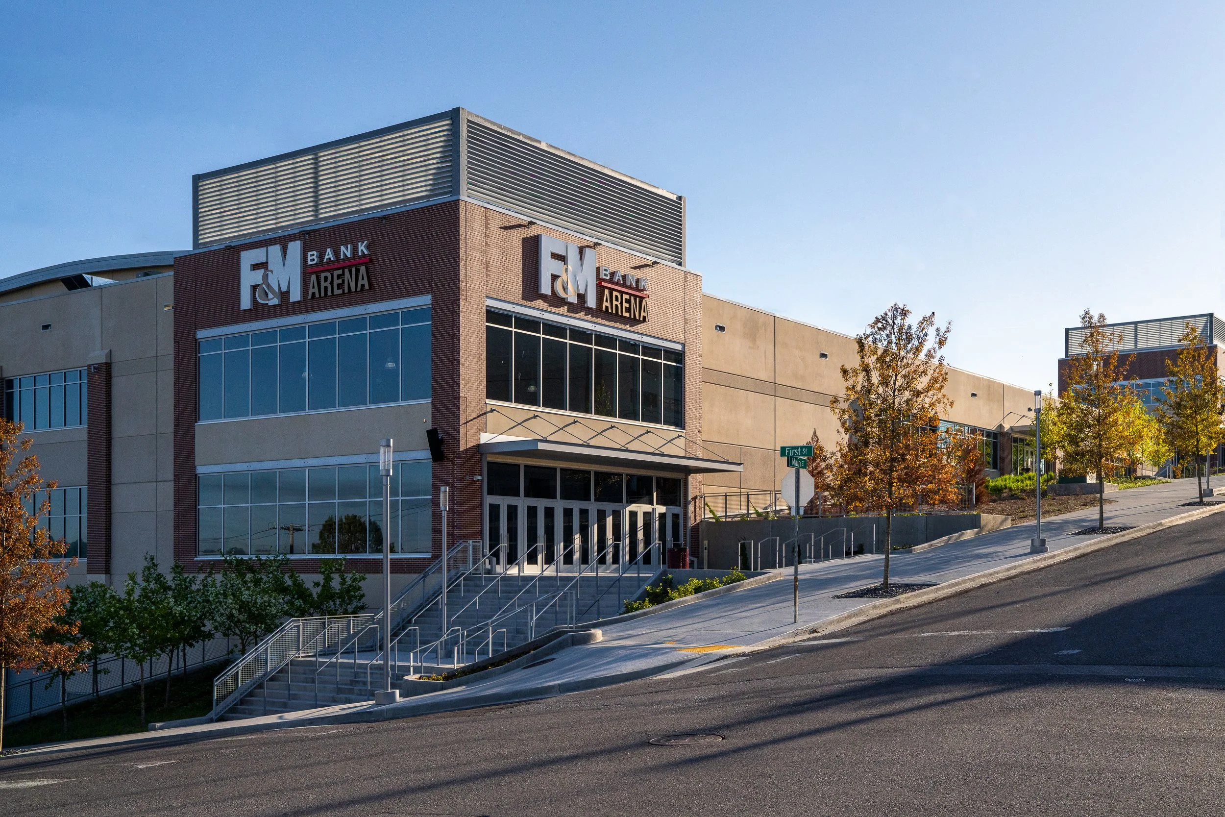 Exterior view of a modern building with the sign 'F&M Bank Arena,' large glass windows, surrounded by trees with fall foliage, and a clear blue sky.