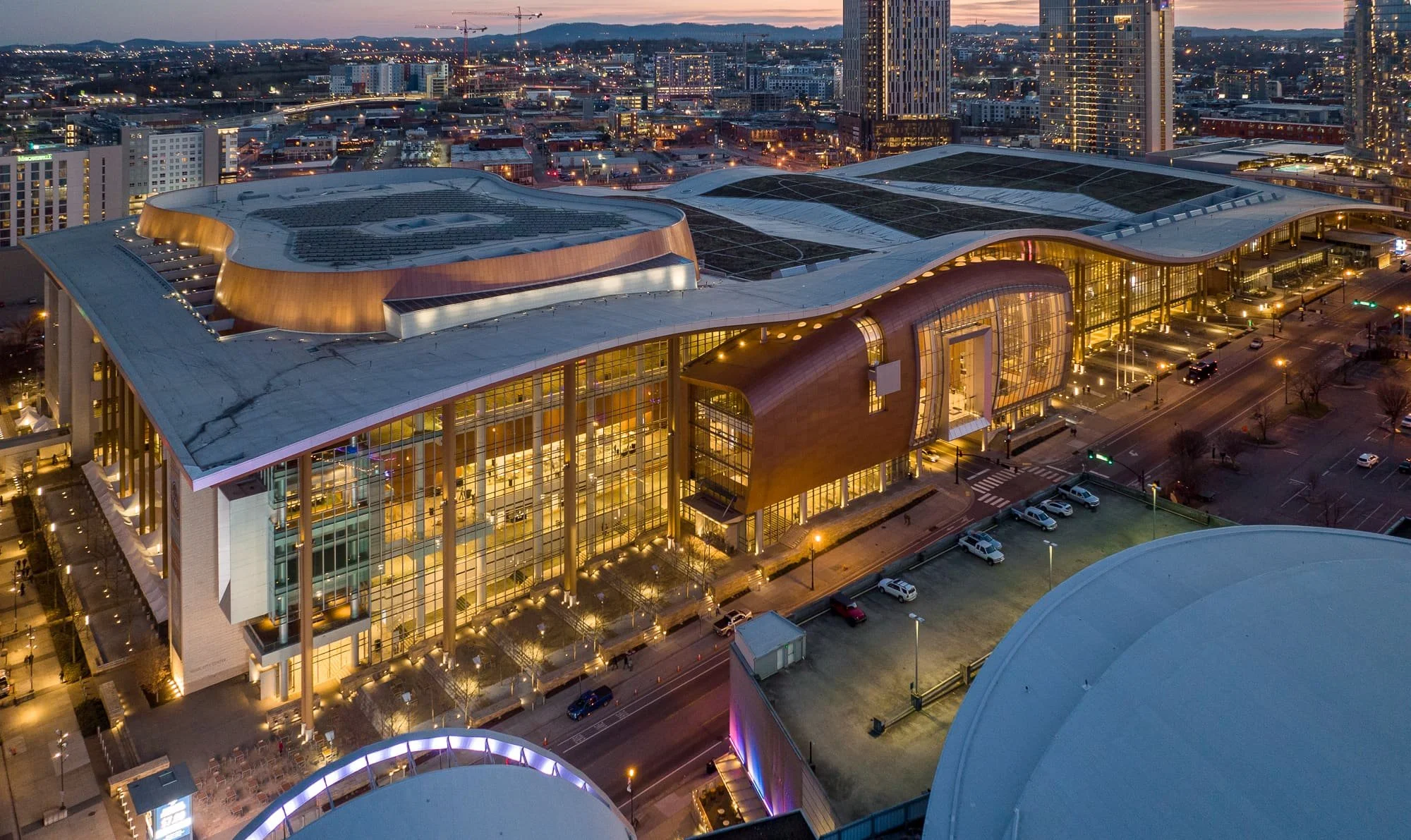 Aerial drone view of Music City Center in downtown Nashville at dusk with its undulating roof, copper facade, illuminated glass curtain wall, green roof sections, and the city skyline beyond