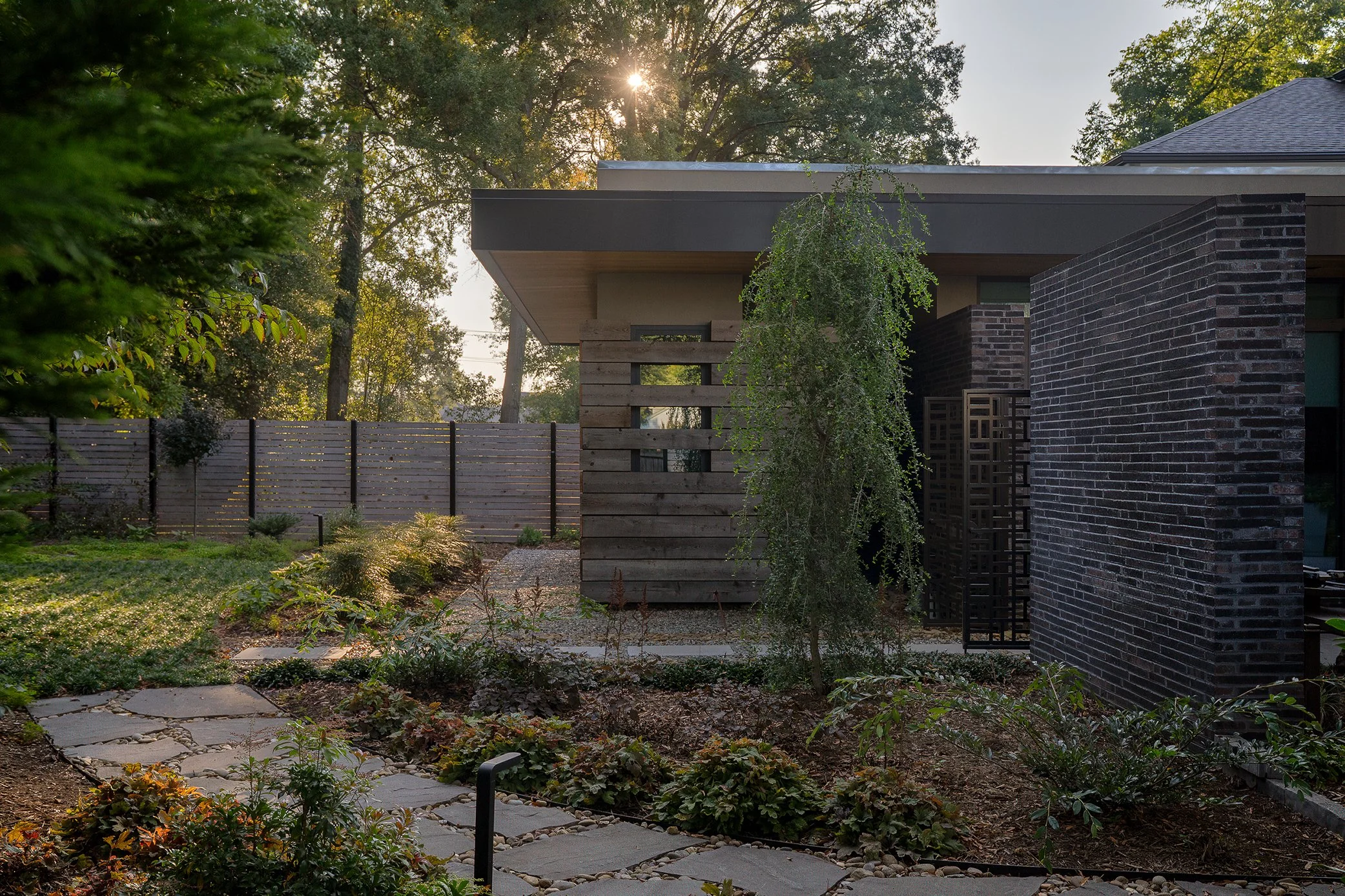 A landscaped garden leads toward a wood entrance screen beneath a broad roof overhang beside a dark brick wall.