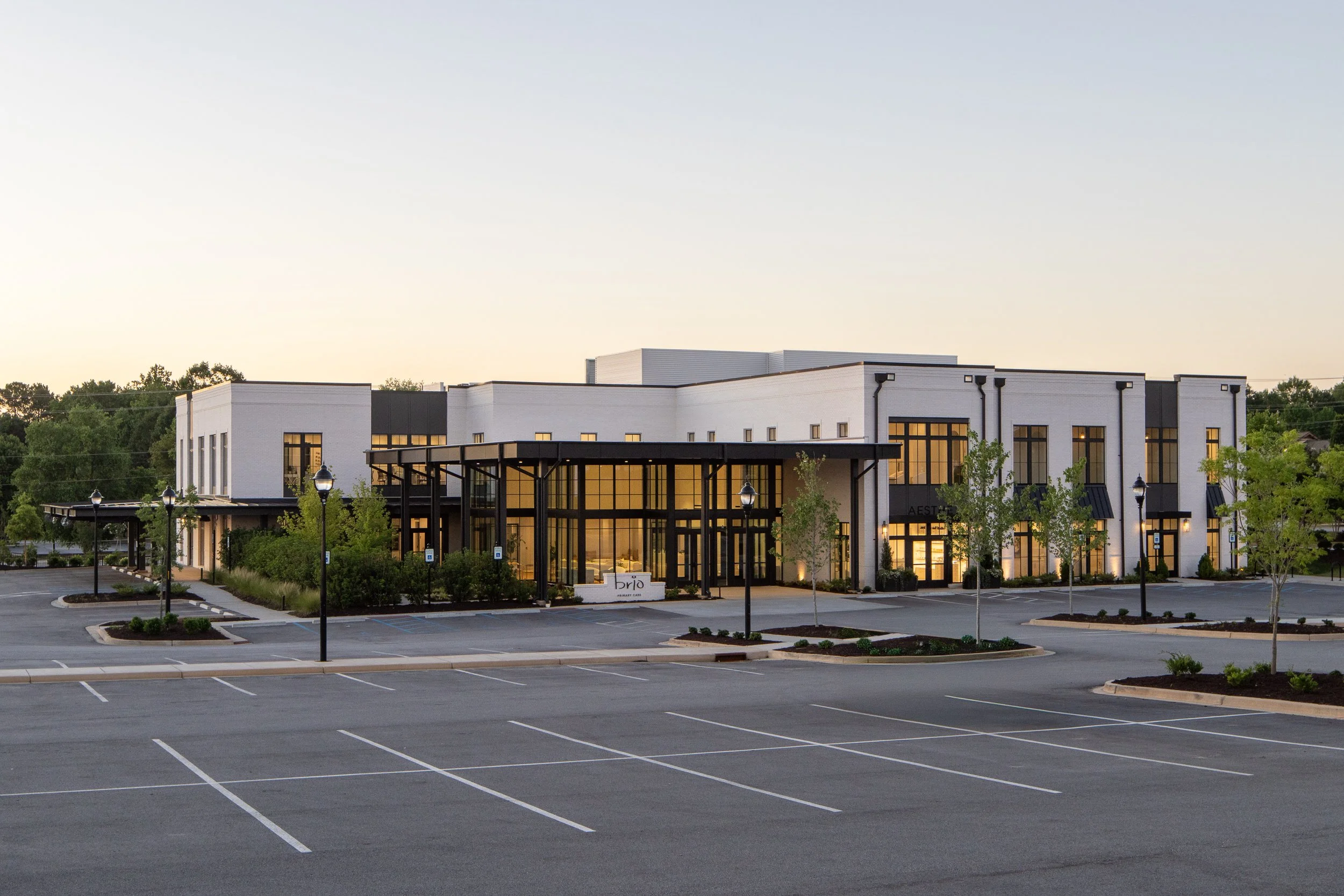 A modern building with large glass windows and a white exterior, surrounded by a parking lot with empty parking spaces and a few small trees. The sky above is clear with a faint gradient from light to darker blue.