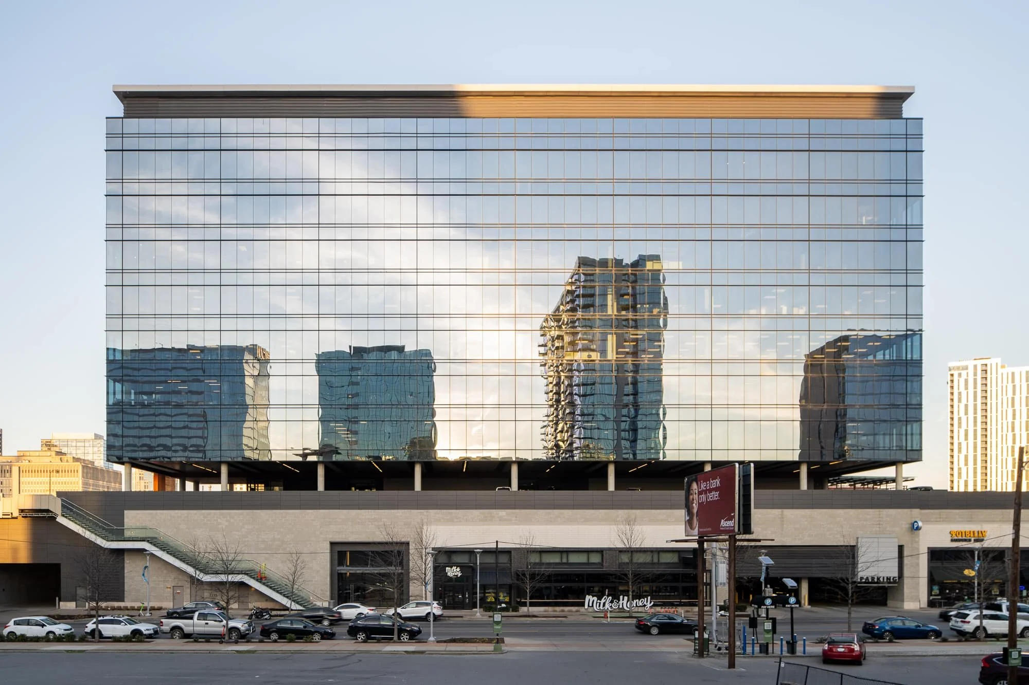 Full facade of Gulch Crossing in Nashville showing the curtain wall glazing reflecting surrounding buildings, raised podium base, street level retail including Milk and Honey, and surrounding Gulch streetscape