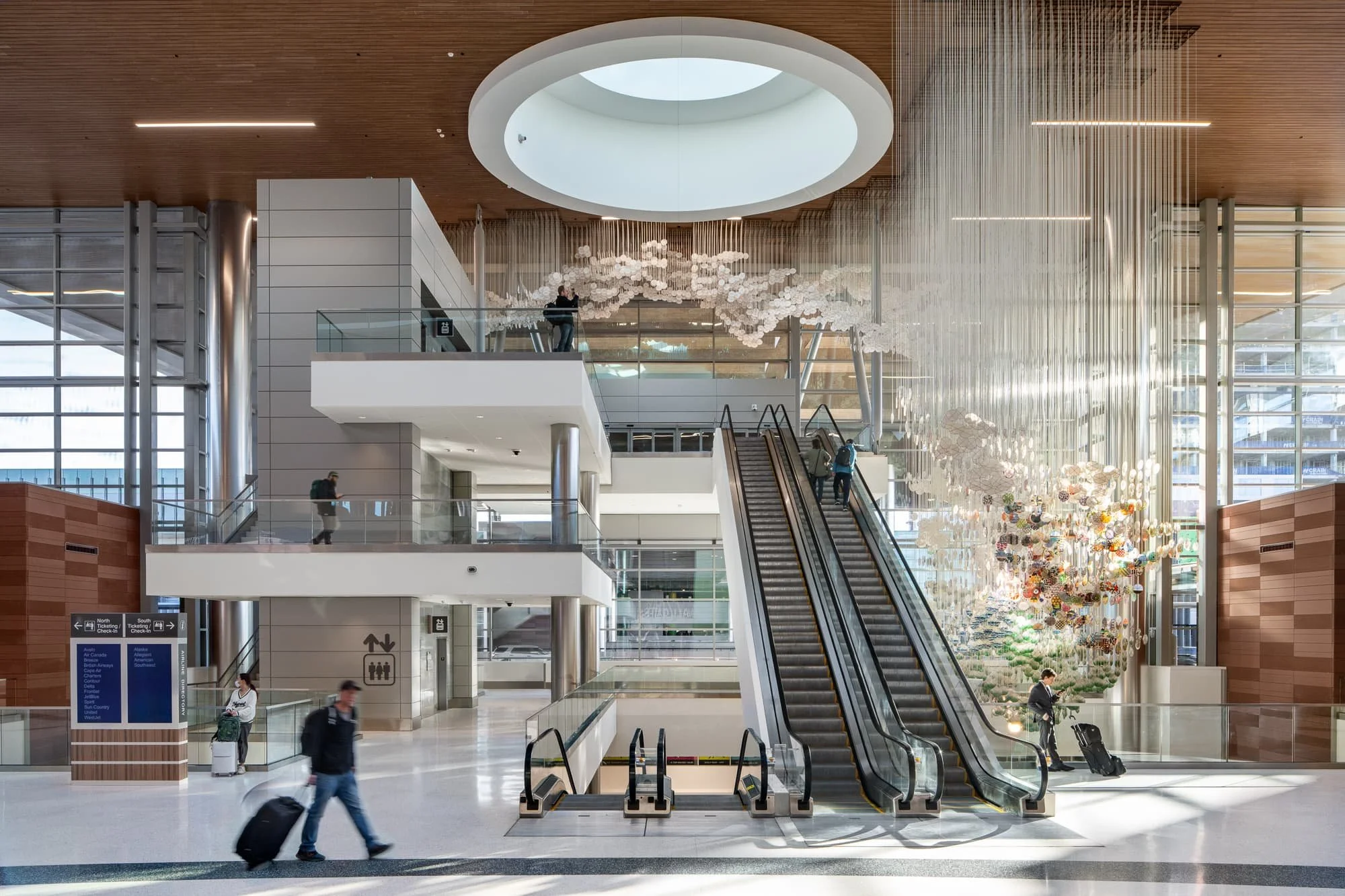 Interior of a modern airport with escalators, large windows, and decorative hanging art fixtures.