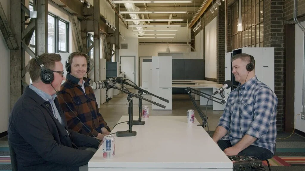 Three men sitting at a table in a recording studio with microphones and headphones, engaged in a conversation for a podcast or radio show.