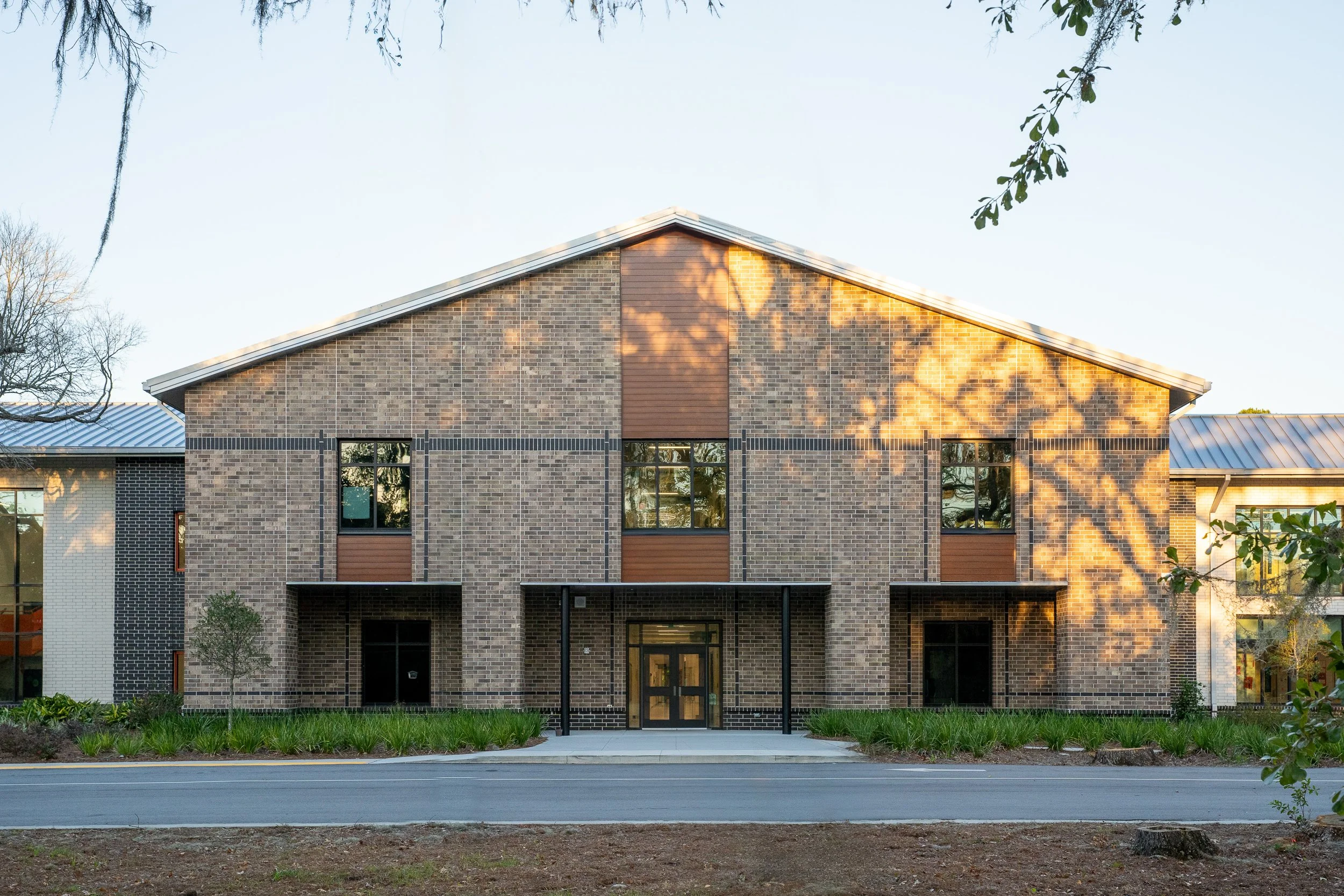 Straight-on view of the gymnasium facade with brick walls, metal roof, black brick banding, and warm tree shadows across the elevation.