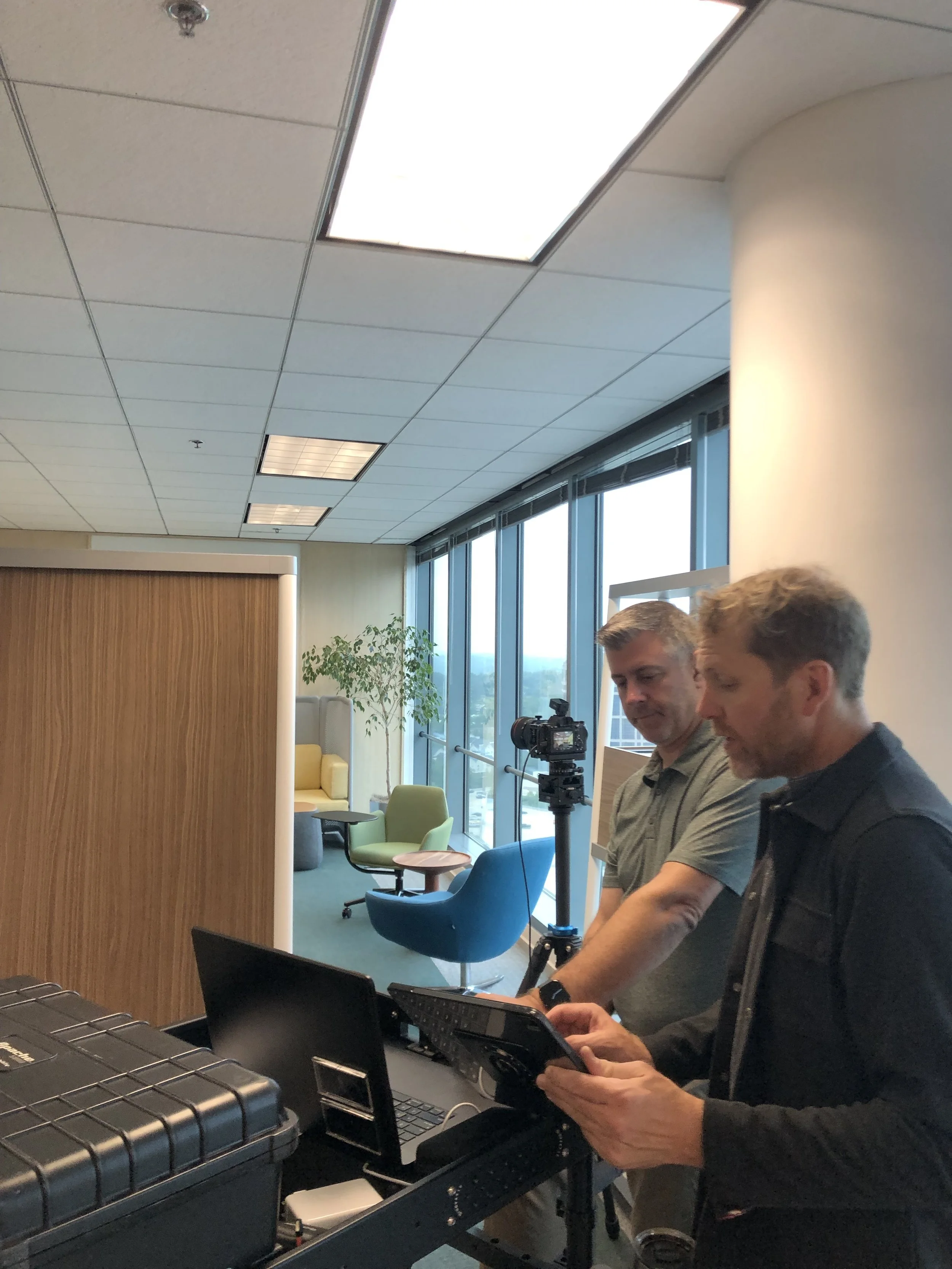 Two men are standing next to each other in an office, working on laptops. One is holding a bar code scanner. The background shows large windows, chairs, and a potted plant.