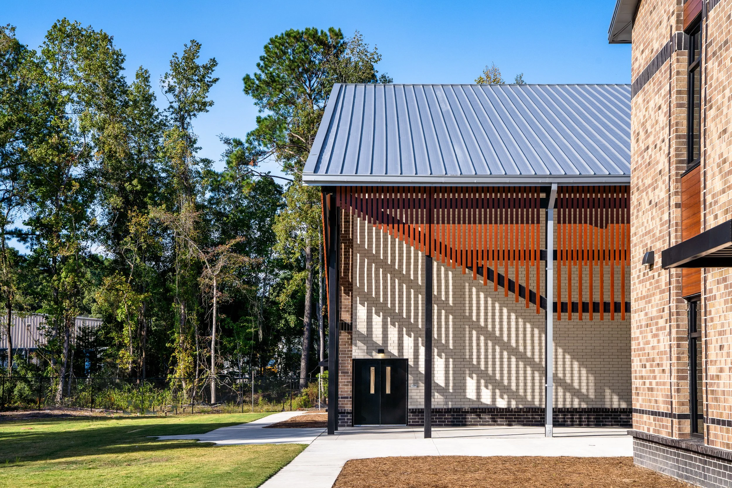 Exterior detail of the brick school building with metal roof, vertical wood screen, and strong afternoon shadows across the facade.
