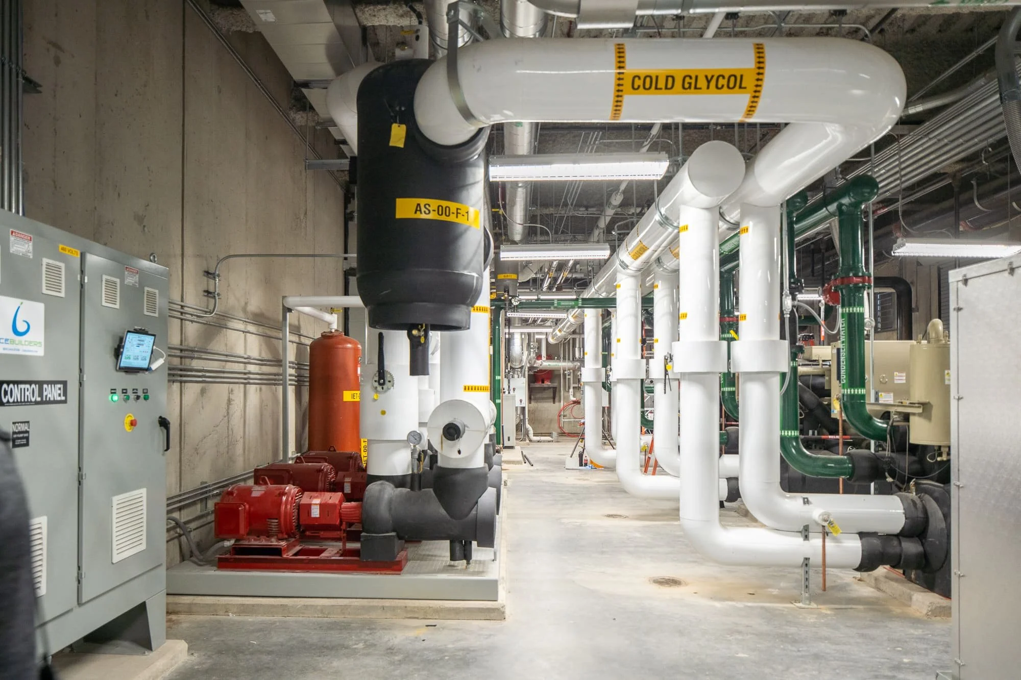 Industrial mechanical room with labeled cold glycol piping, red pumps, control panels, and exposed overhead utility systems.