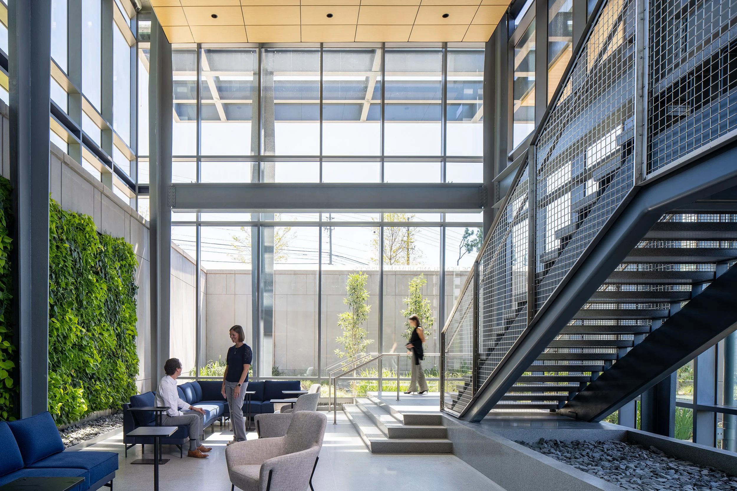 Double-height atrium with a full-height living wall on the left, blue and gray lounge seating, an exposed steel stair on the right, and daylight pouring through the curtain wall to a courtyard beyond.