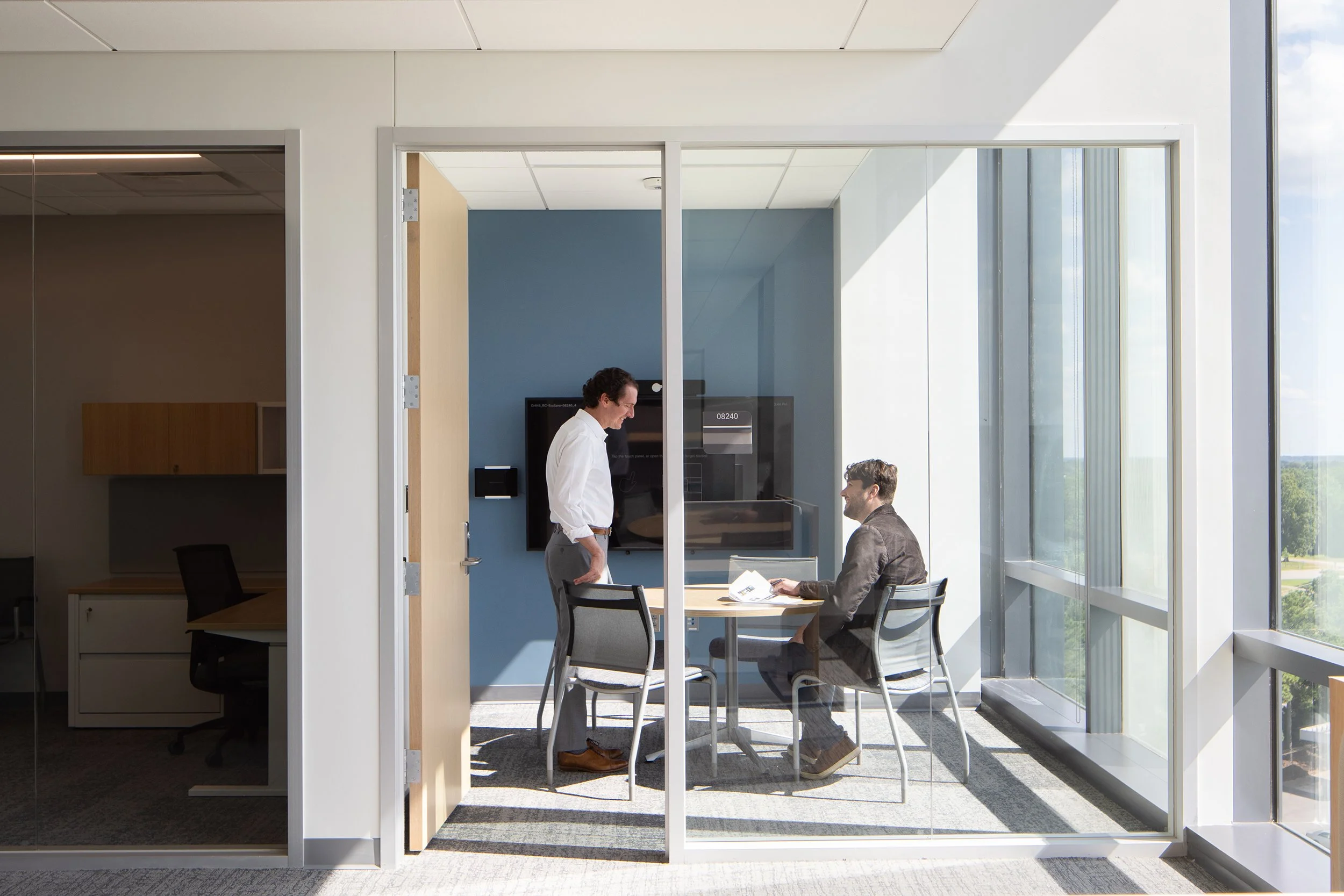 Small glass-walled meeting room with a blue accent wall, two people in conversation around a round table, and a window framing the wooded landscape beyond.