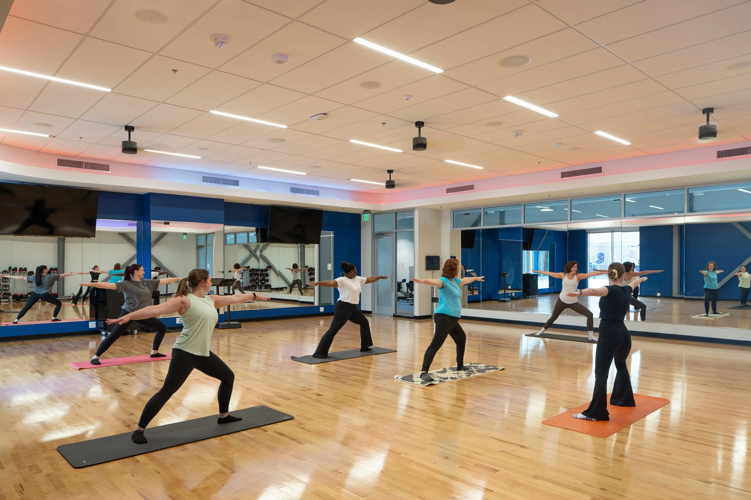 Group exercise class in a mirrored studio with wood floors, yoga mats and soft perimeter lighting.