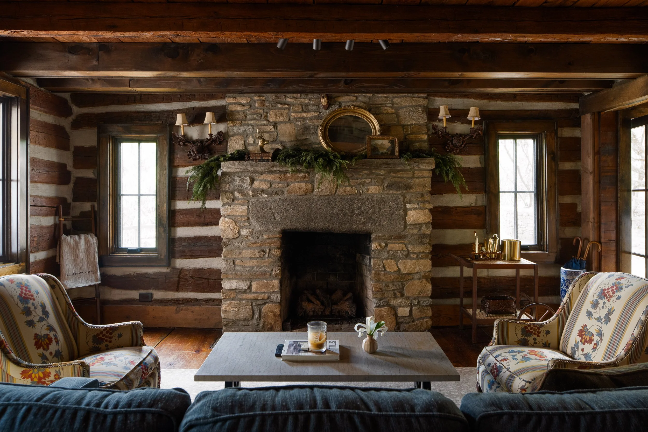 Sitting room with a large stone fireplace, log walls, floral chairs, mantel greenery, and soft window light.