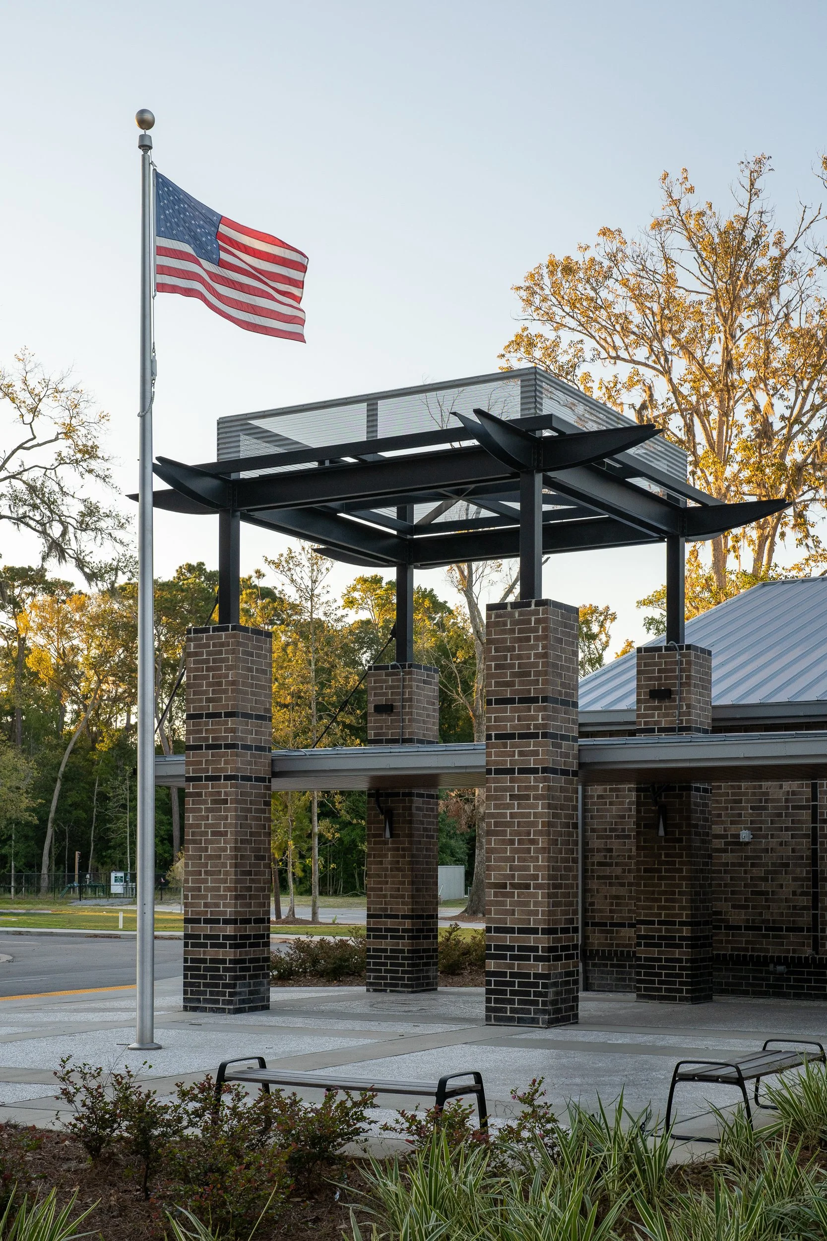 Exterior view of the school entry canopy with brick columns, flagpole, landscaping, and warm morning light on the surrounding trees.
