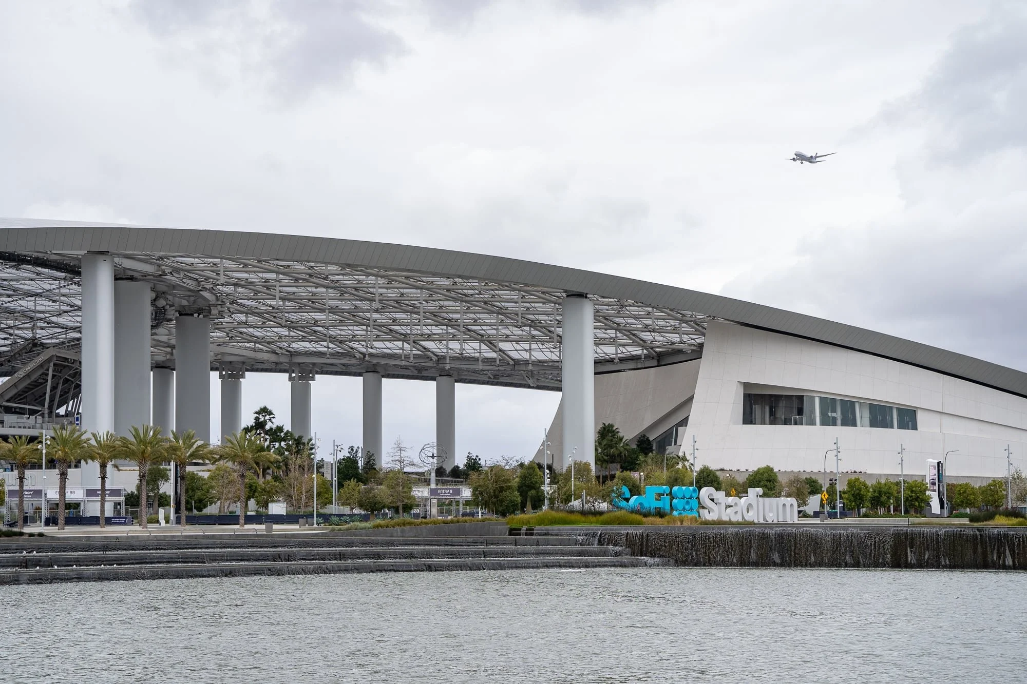 Wide view of a modern stadium across a curved waterfront plaza with canopy roof, columns, and landscaped grounds.