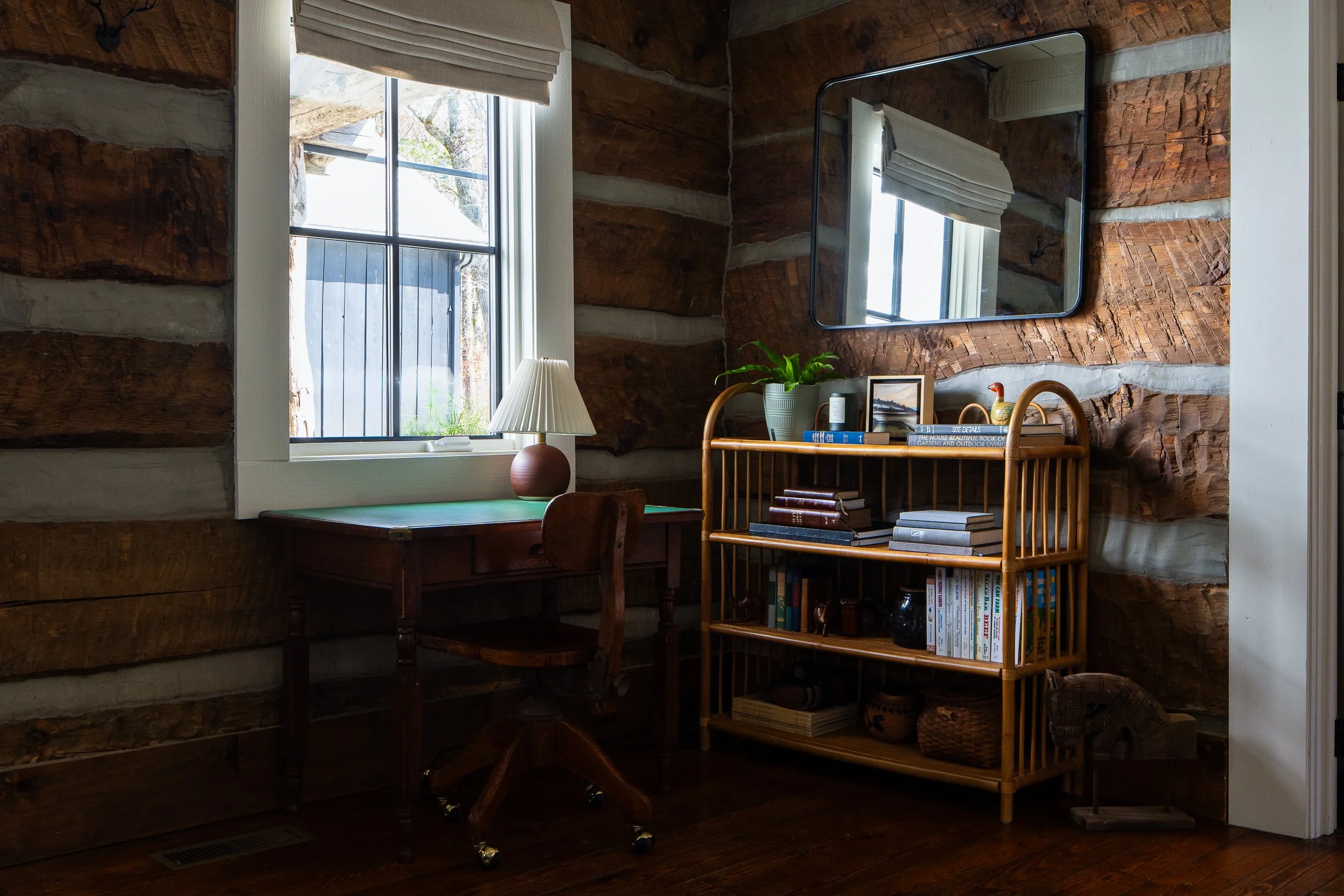 Writing desk and rattan bookshelf set against rustic log walls beside a tall window.