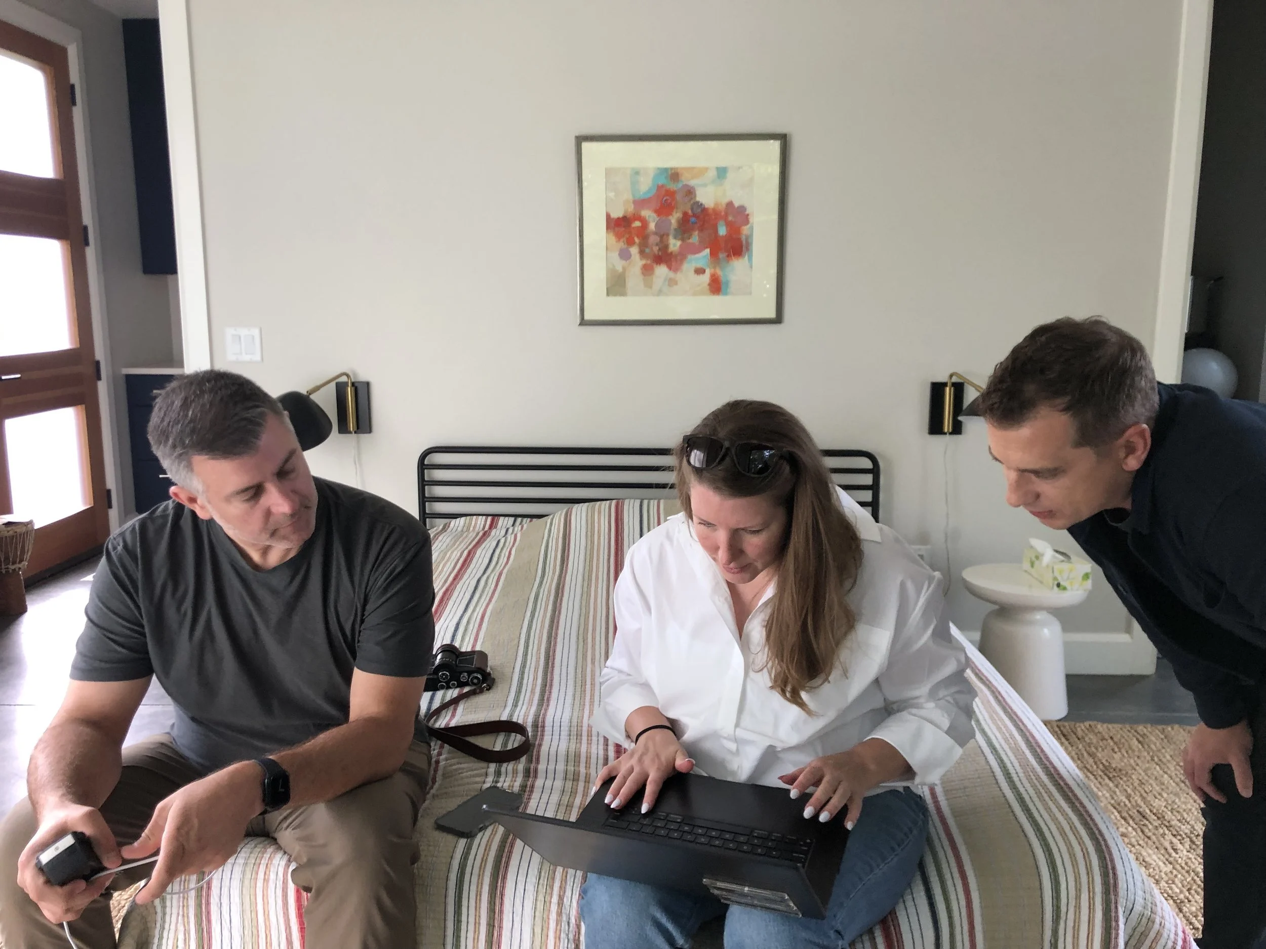 Four people sitting on a bed, looking at a laptop and smartphones, with a painting hanging on the wall behind them.