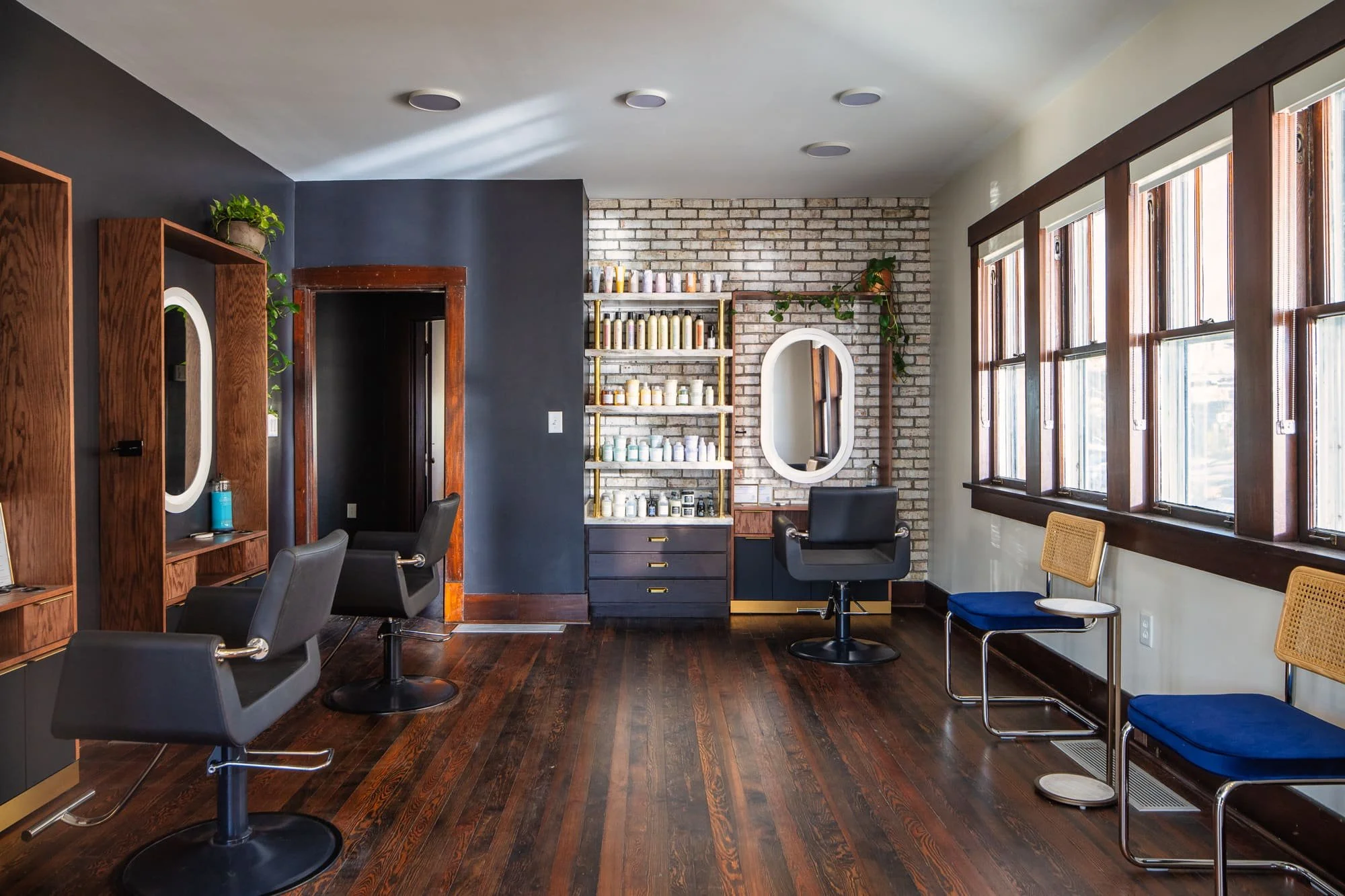 Salon styling area with exposed brick wall, black salon chairs, round mirror, product shelving, and warm wood floors.