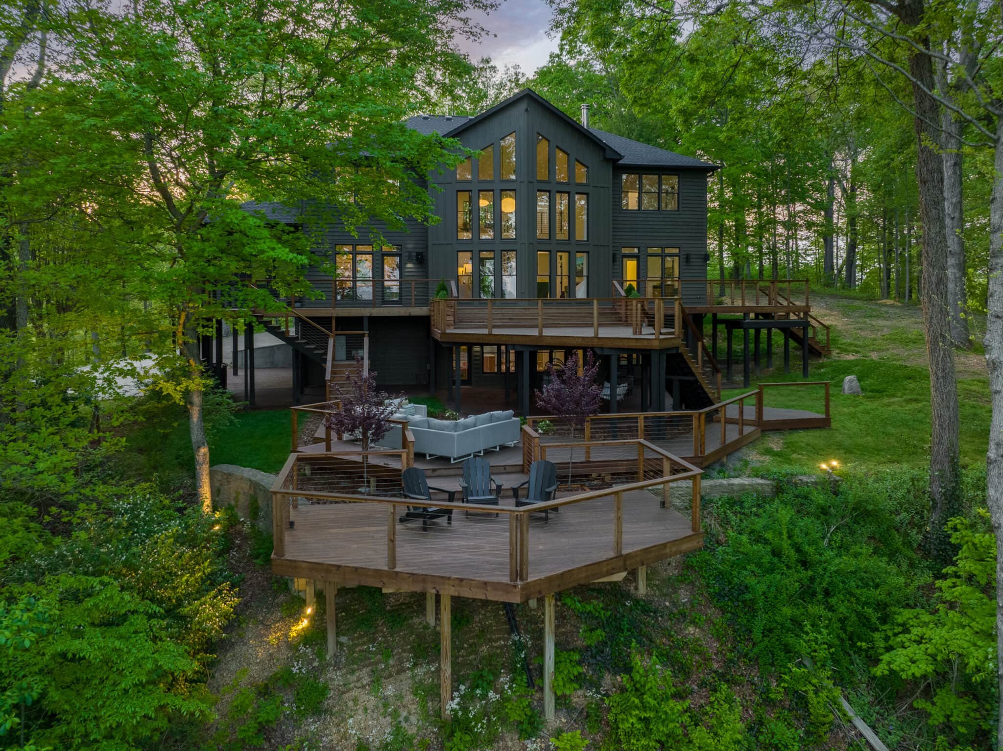 Rear view of a large dark-sided home with expansive wood decks, warm interior lights, and surrounding trees.
