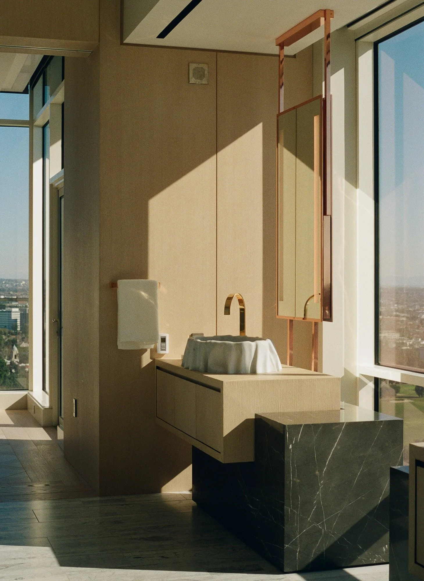Sunlit high-rise bathroom with a sculptural sink, copper mirror, black marble base, and expansive city views.
