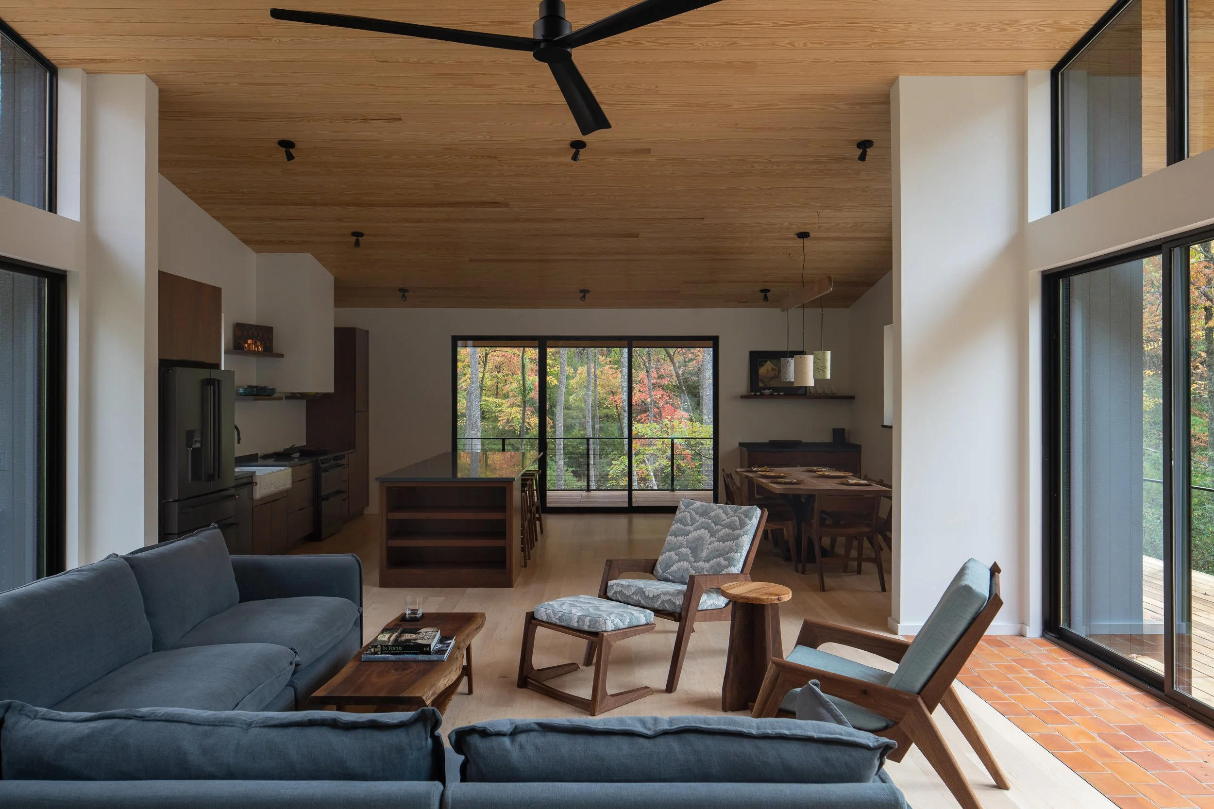 Open living, dining, and kitchen area with wood ceilings, large windows, and views into the autumn forest.