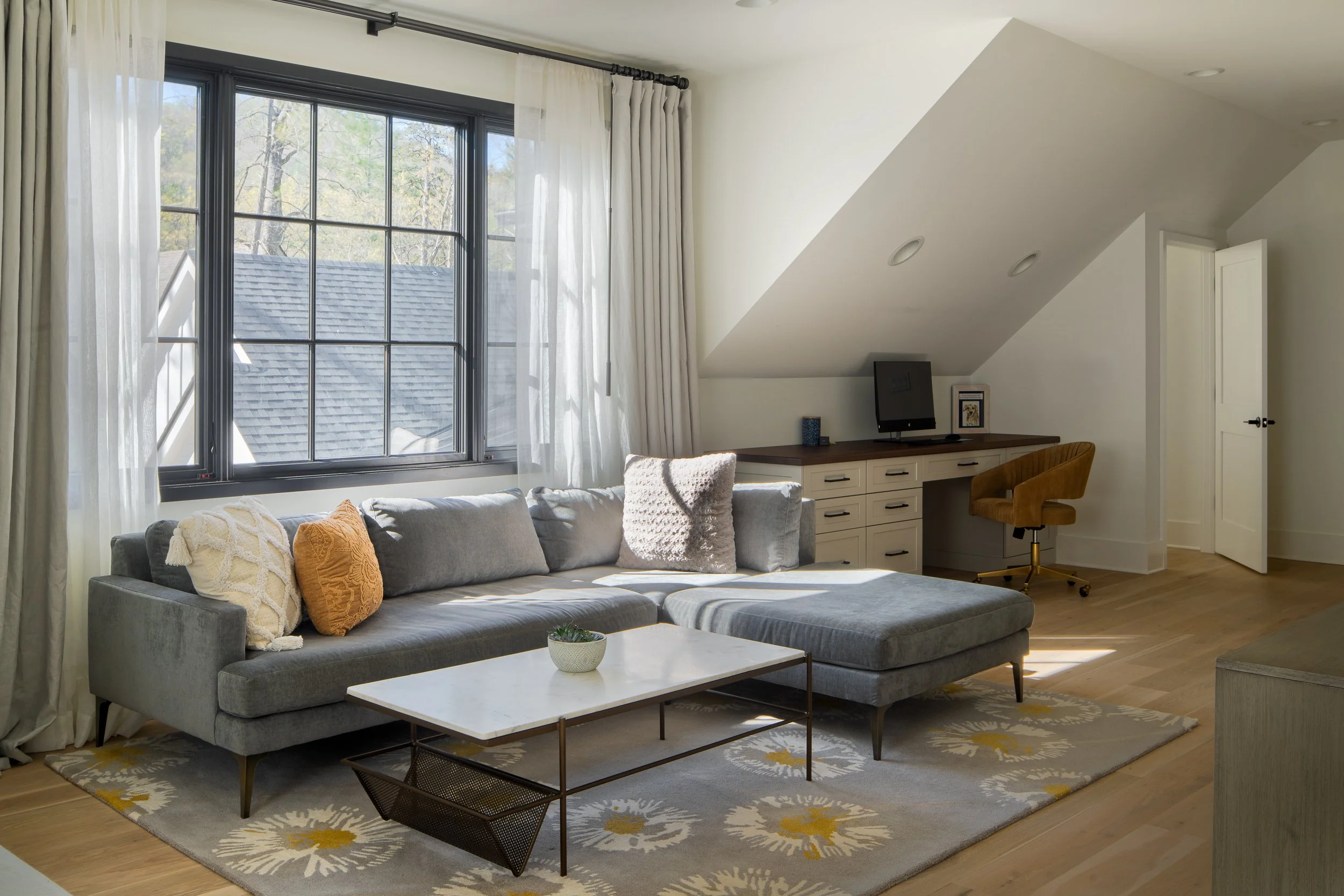 Home office with a sectional sofa and built-in desk beneath a sloped ceiling.