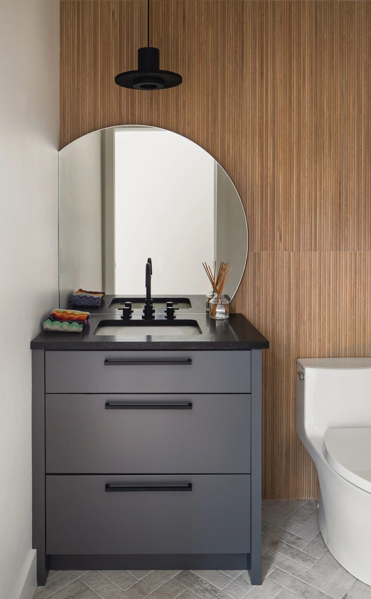 Powder room with a gray vanity, round mirror, black fixtures, and vertical wood slat wall.