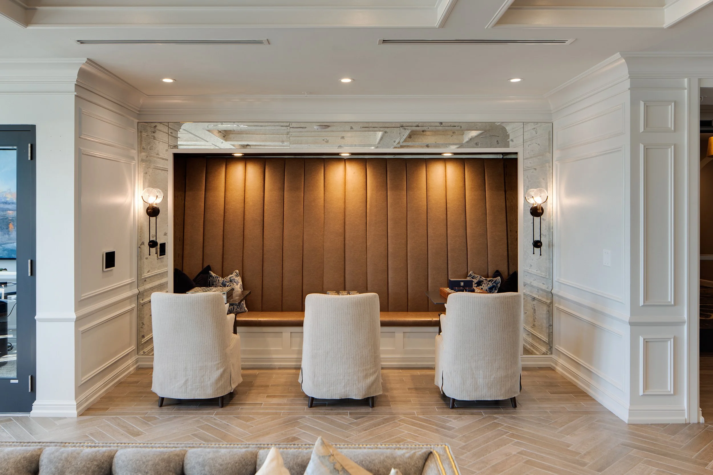 Intimate lounge niche with a brown leather channel-tufted banquette, antiqued mirror surround, paired wall sconces, three slipcovered armchairs, and a herringbone wood floor.