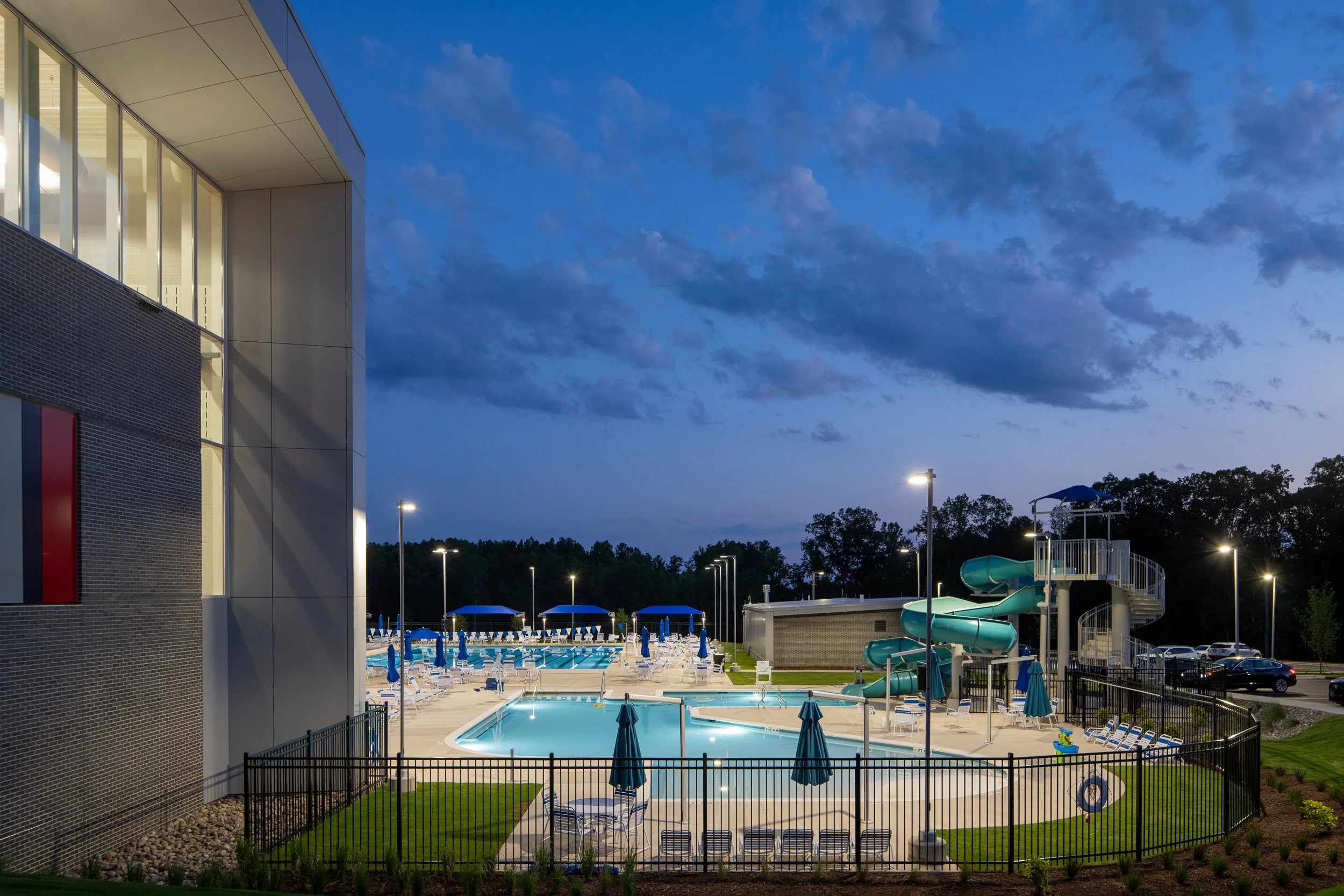 Evening pool deck with waterslides, lounge chairs, umbrellas and illuminated paths beside the YMCA building.