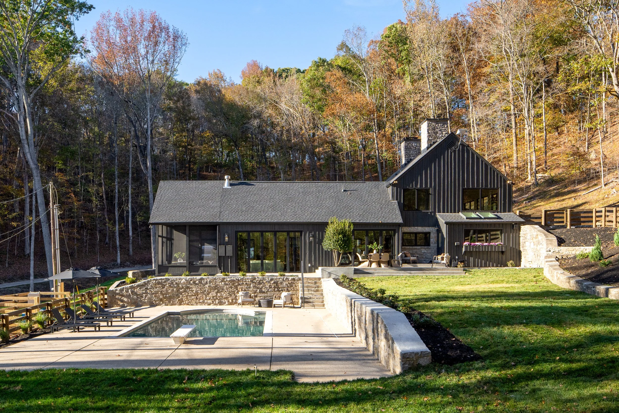A modern house with black vertical siding, large windows, and a gray roof, situated in a scenic area with a wooded hillside in the background and a backyard with a swimming pool and patio furniture.