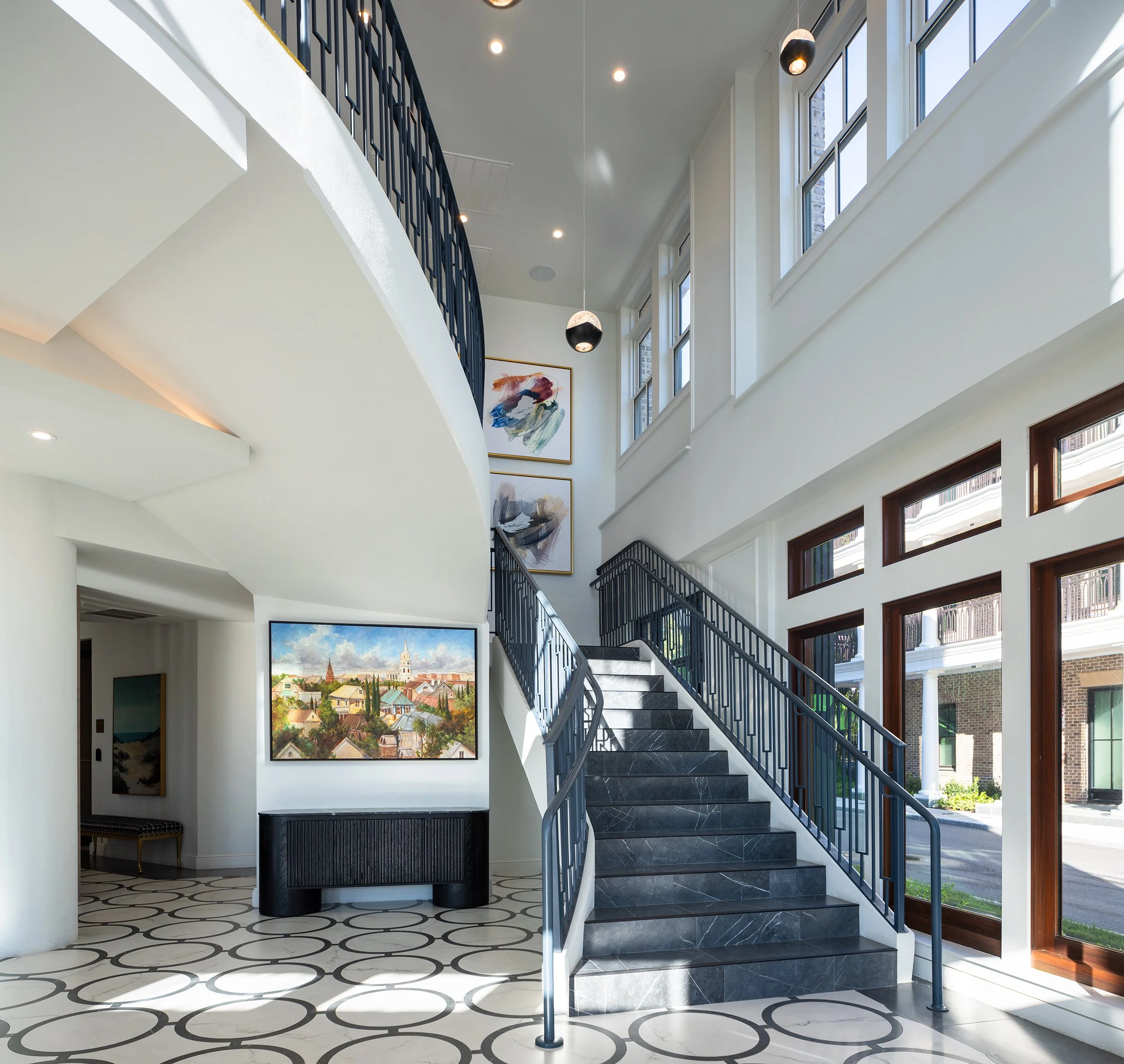 Grand interior stair with dark marble treads, a dark blue iron railing in a geometric pattern, large windows with mahogany trim, paired pendant globe lights, and a Charleston cityscape painting on the wall above the landing.